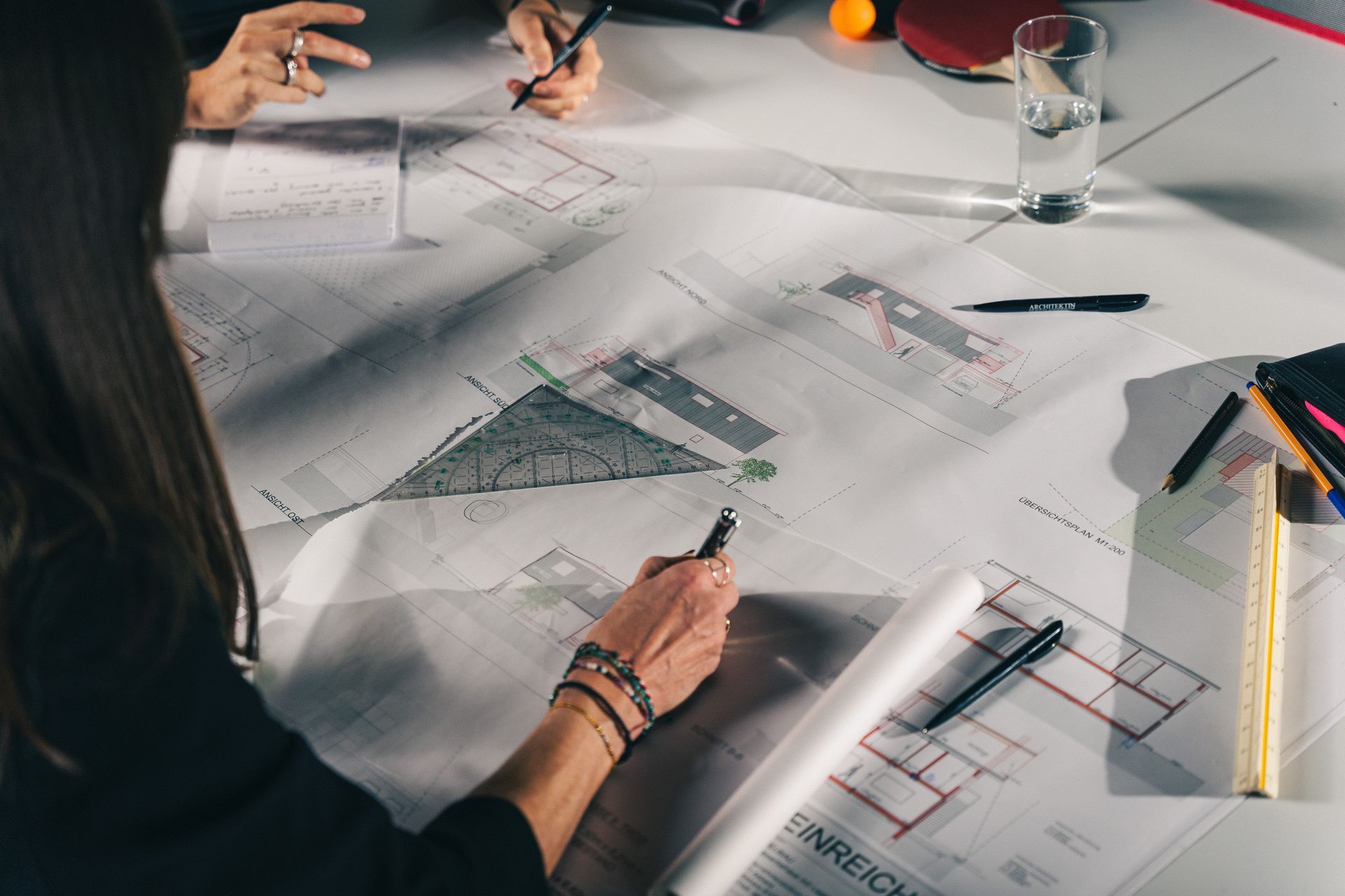 People reviewing architectural plans on a table, with pens, papers, and drafting tools spread out.