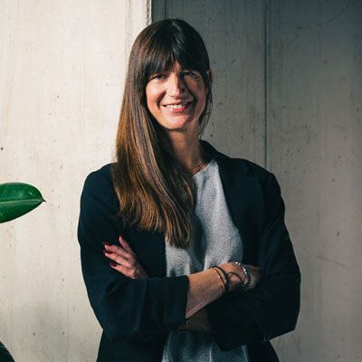 Smiling woman with crossed arms, long brown hair, and dark blazer standing against a concrete wall.