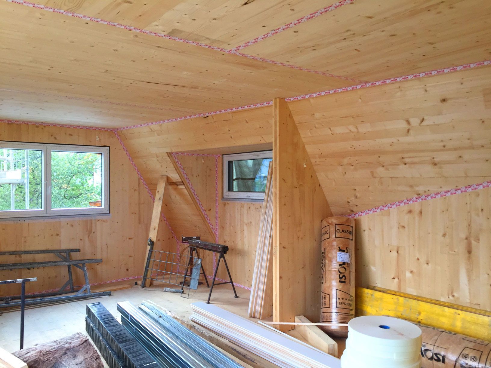 Wood-paneled room under construction with stacked lumber, a ladder, and windows letting in daylight
