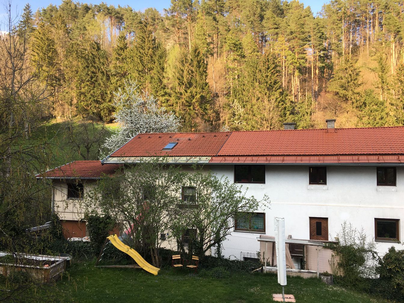 White house with red roof and yellow slide in a wooded yard, with forested hills in the background
