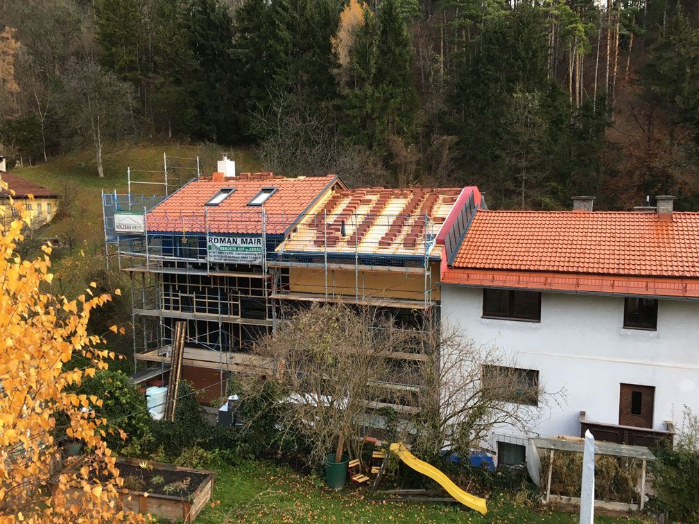 Houses with orange-tiled roofs, one under construction with scaffolding, beside trees on a hillside.