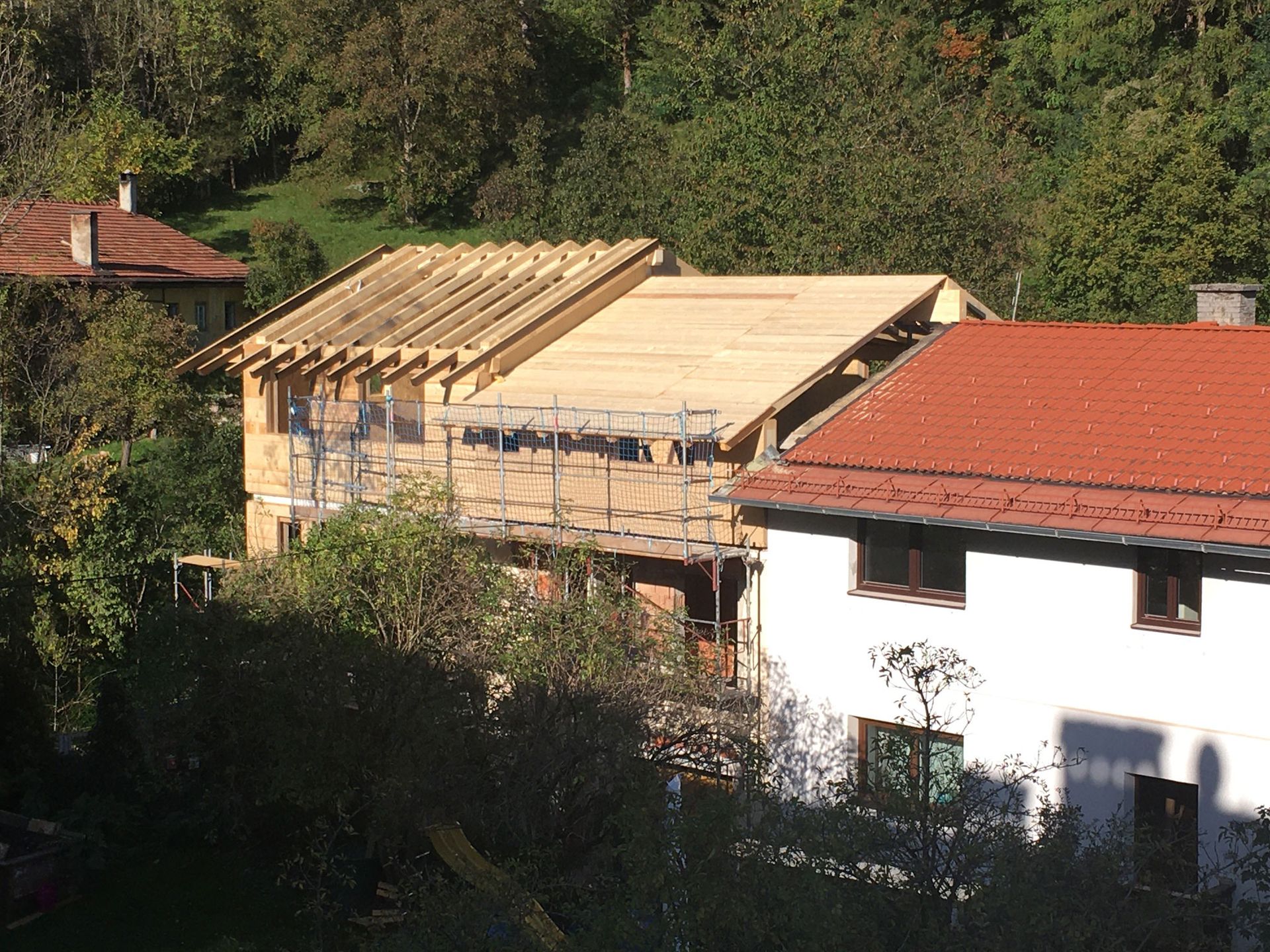 House under construction beside a white home, with a red roof and trees in the background