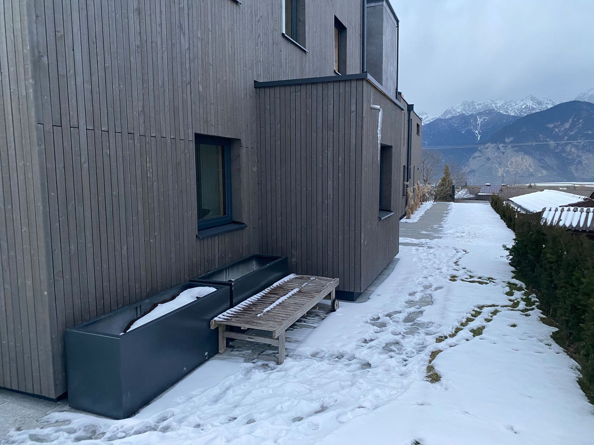 Snowy patio beside gray building with a bench, planters, and mountains in the distance