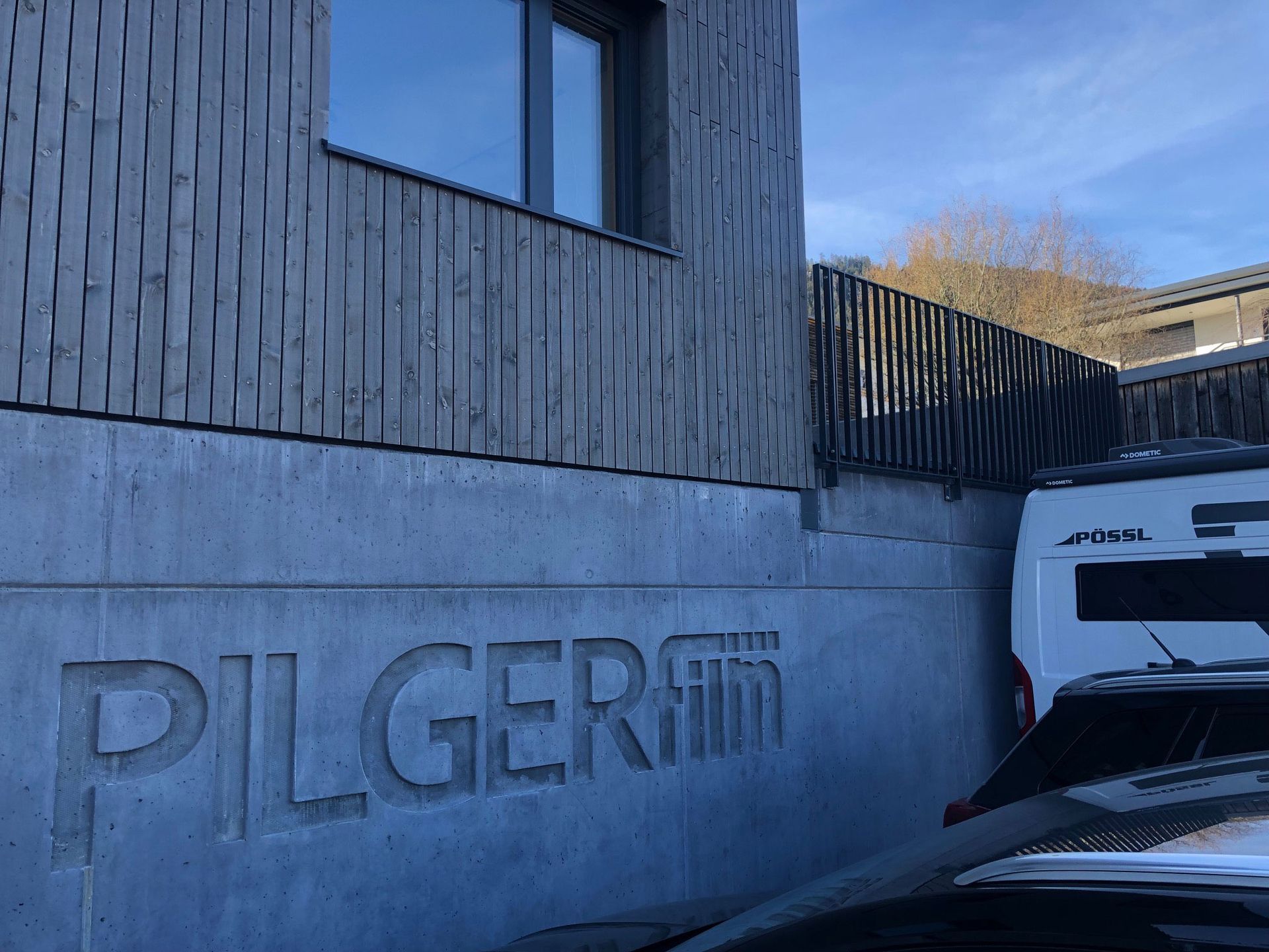 Concrete building wall with “PILGER” lettering, blue sky, and parked vehicles beside it