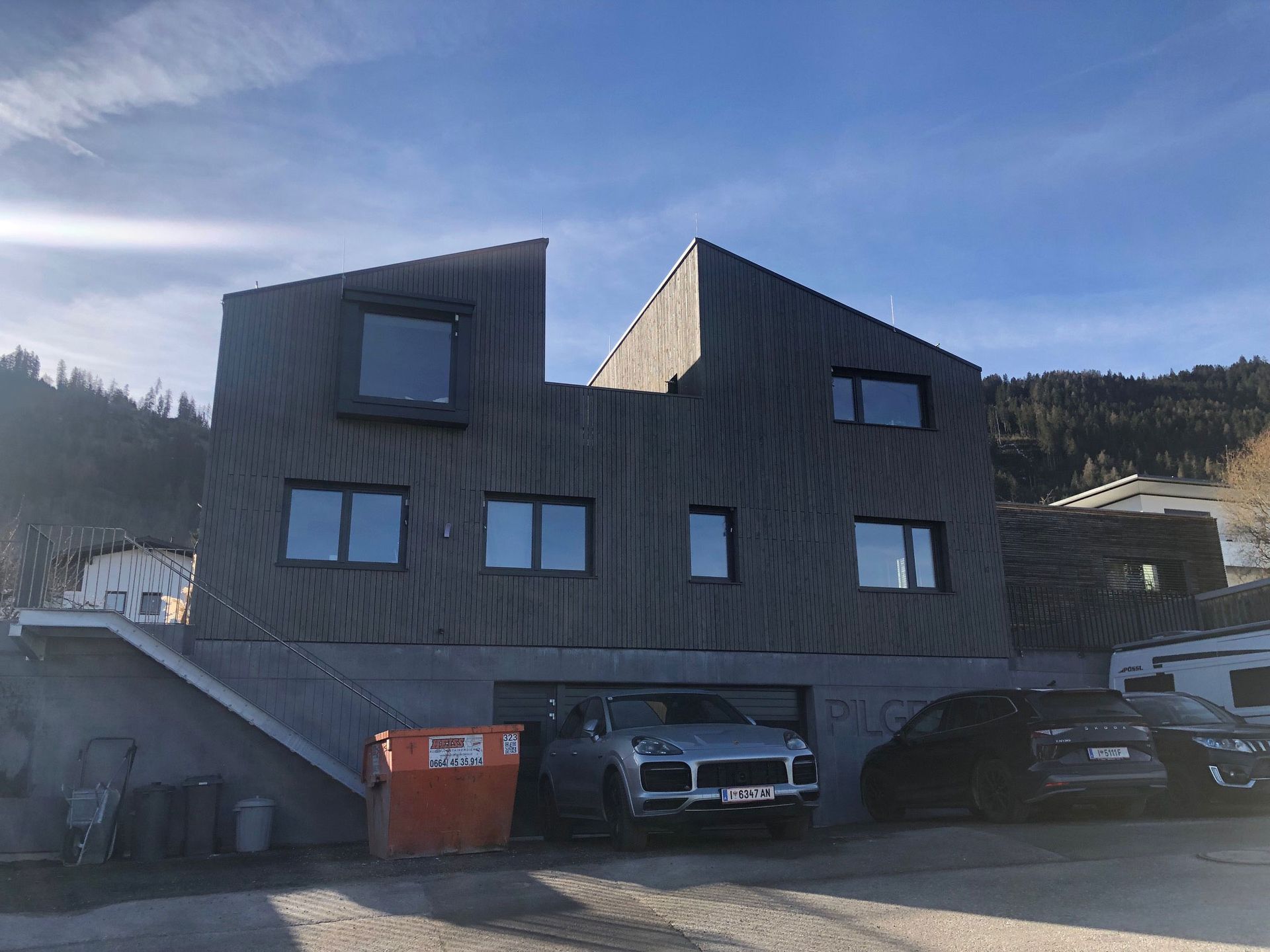 Modern gray house with angular roofs and parked cars in front, under a blue sky
