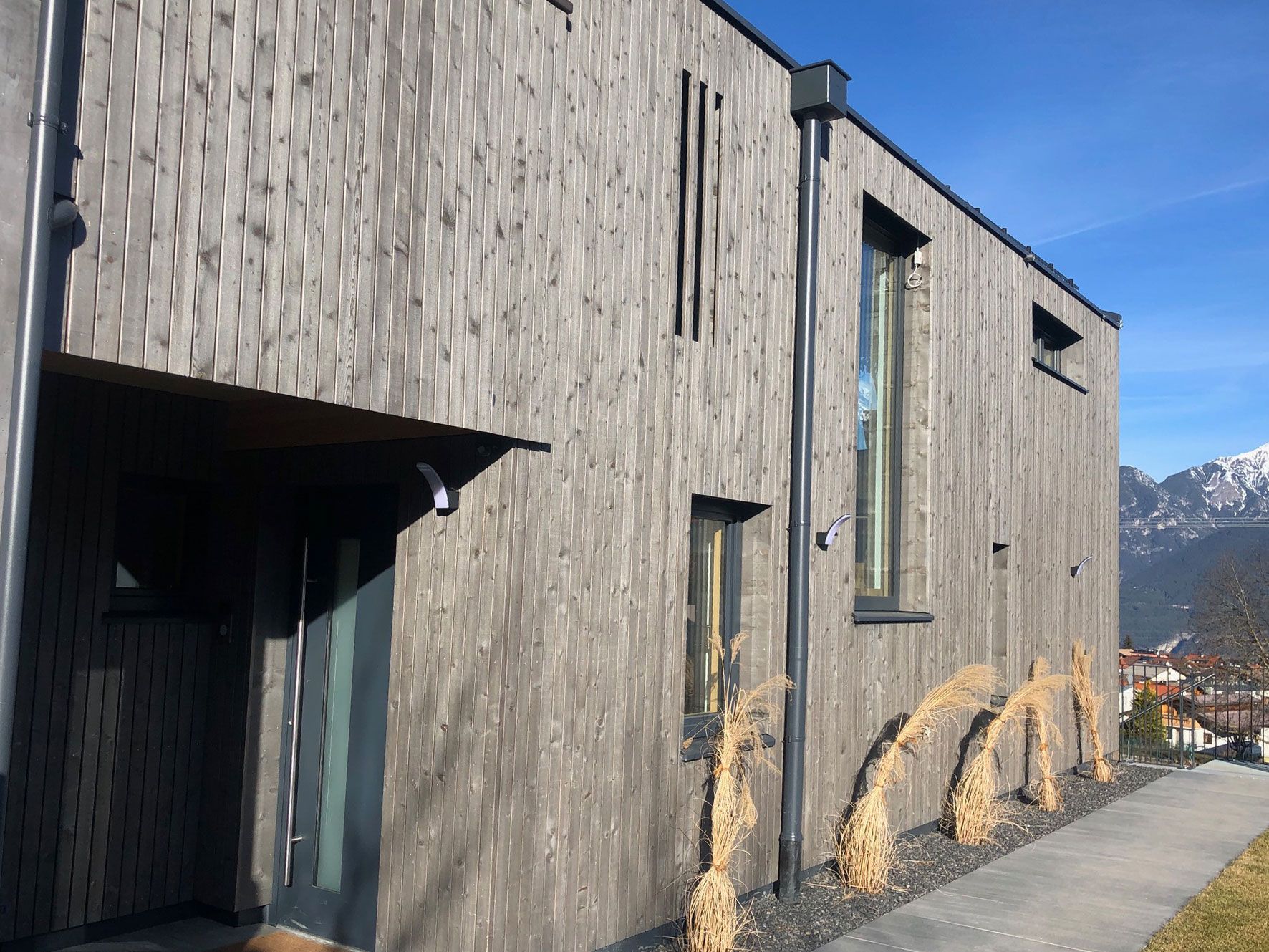 Modern gray wood-sided building with small windows beside a grassy path under a blue sky.