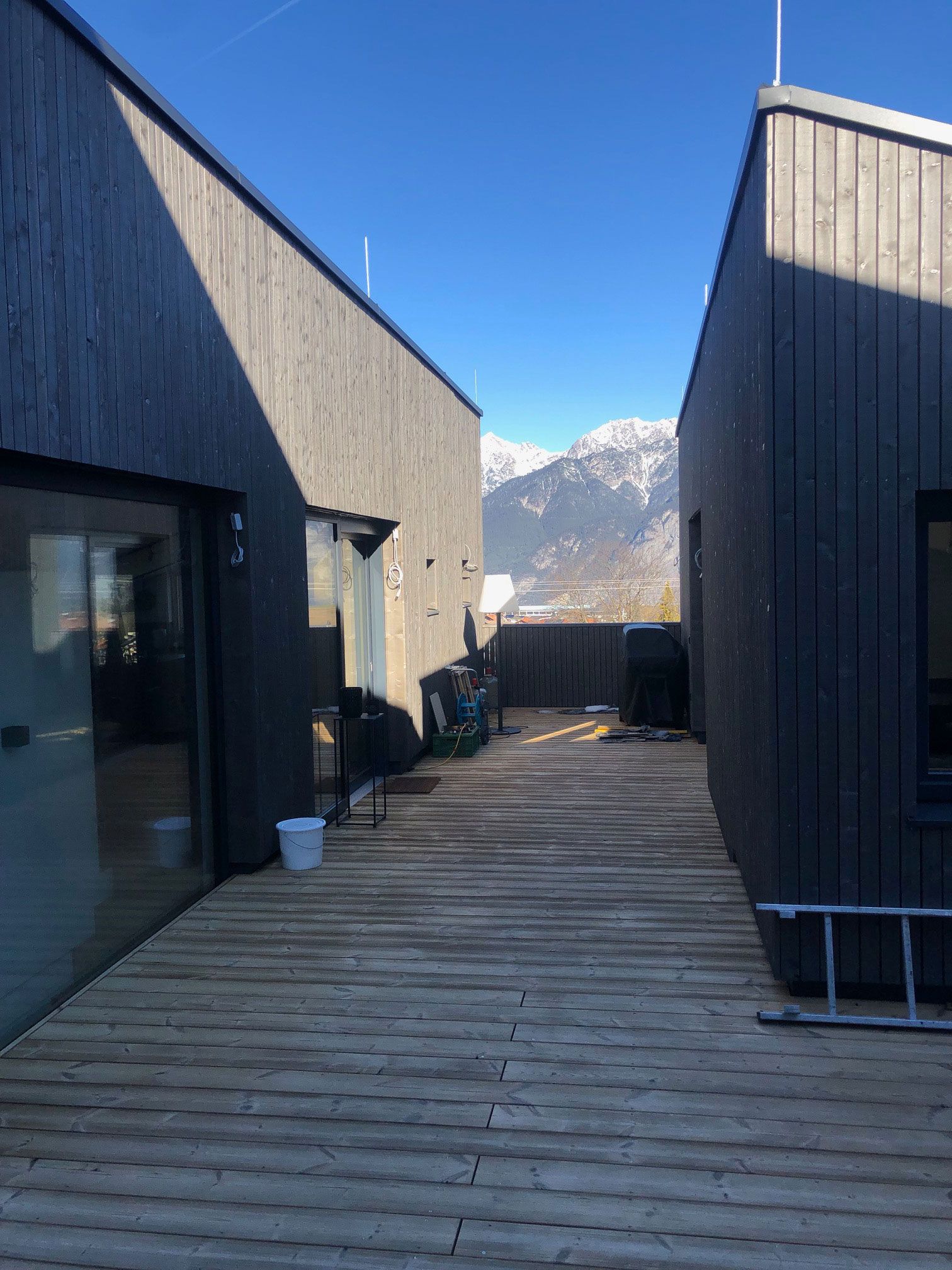 Wooden deck between dark modern buildings, with snowy mountains in the distance under a blue sky.