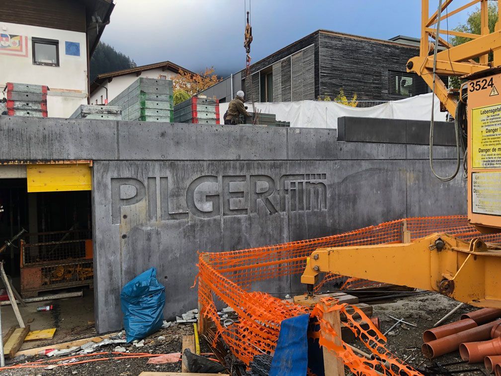 Construction site with concrete wall reading “PILGERMO,” orange safety netting, and yellow machinery