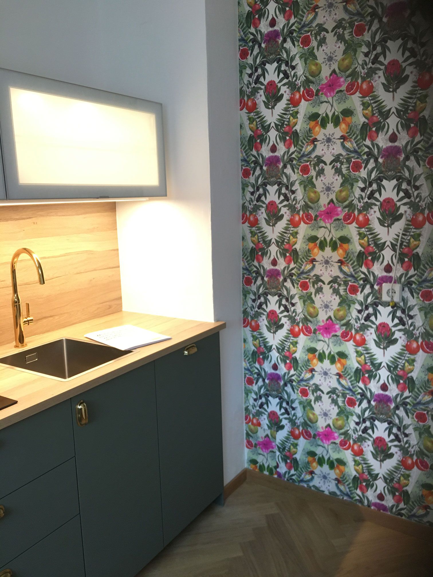 Modern kitchen corner with dark cabinets, sink, lit backsplash, and colorful floral curtain.