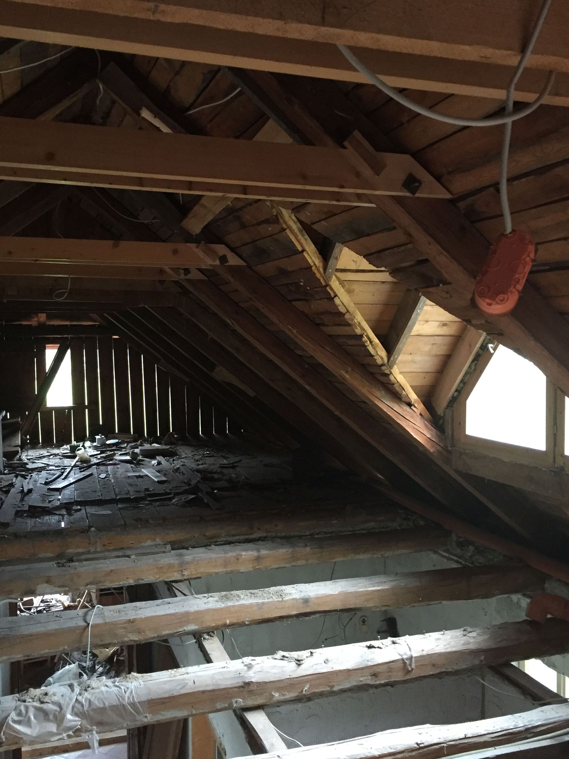 Interior of a damaged attic with exposed wooden beams, debris, and sunlight through small windows