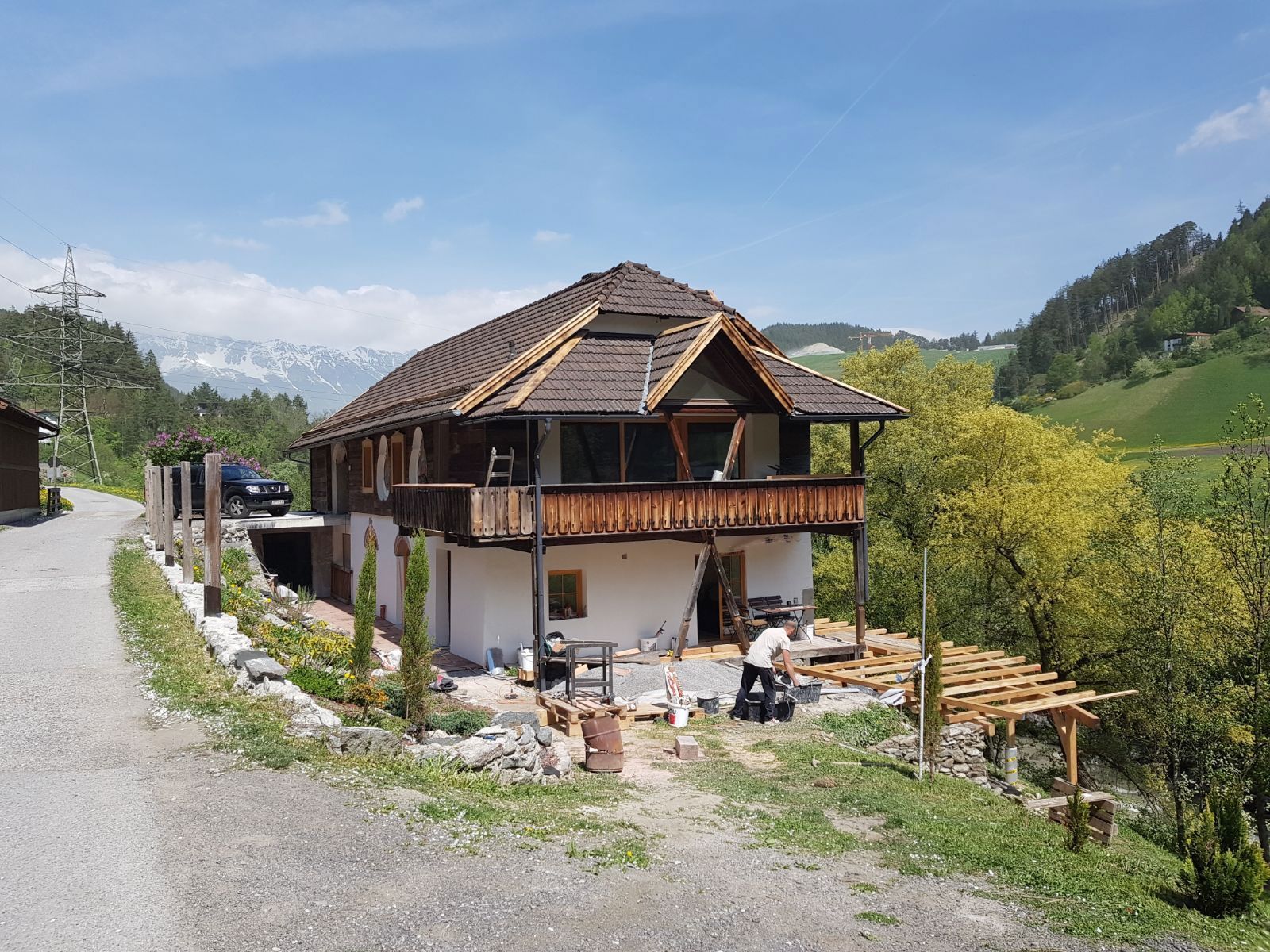 Rural house with wooden balcony beside a gravel road, surrounded by green hills and trees under a blue sky.
