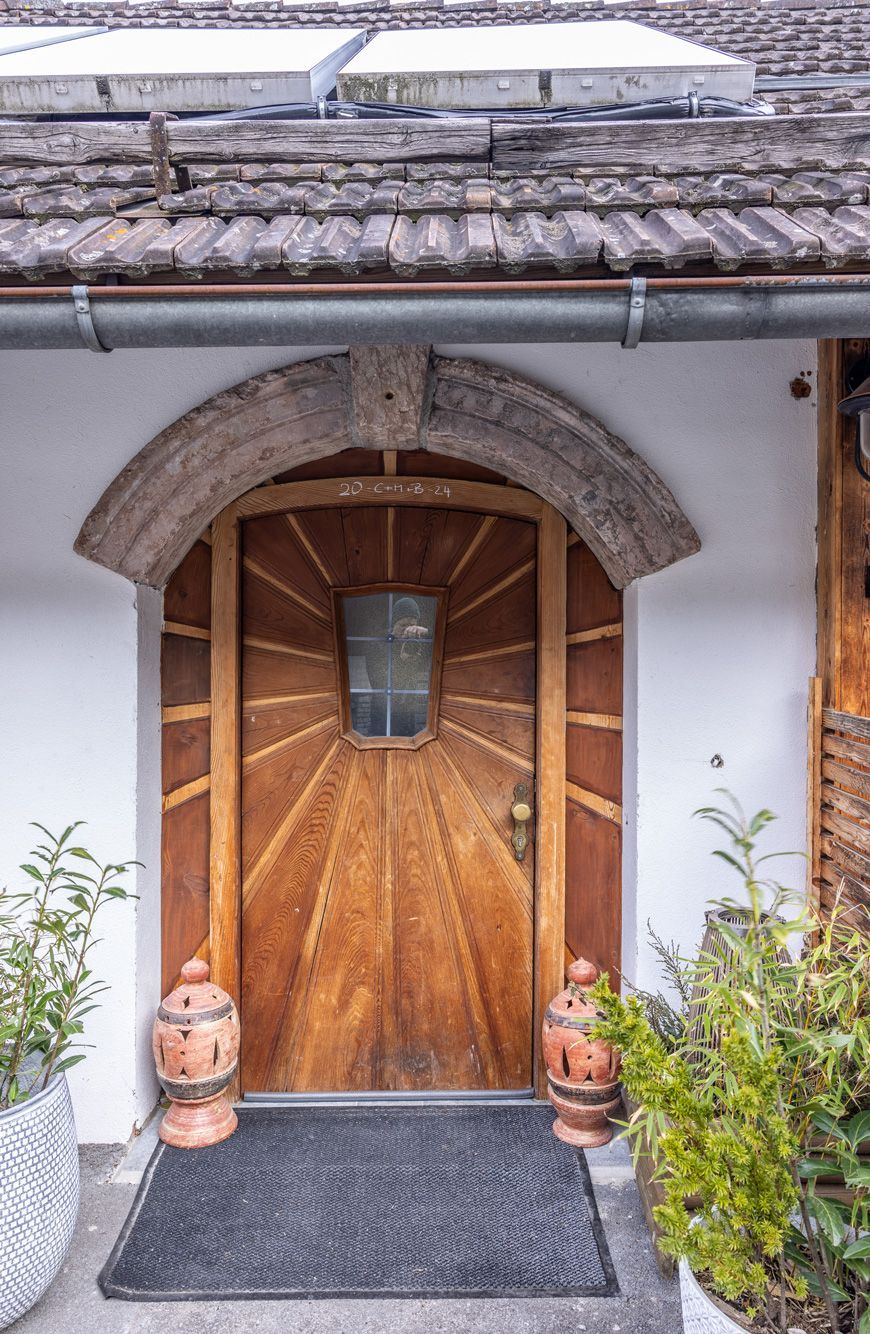 Wooden arched front door with stone trim, potted plants, and a dark doormat under a tiled awning