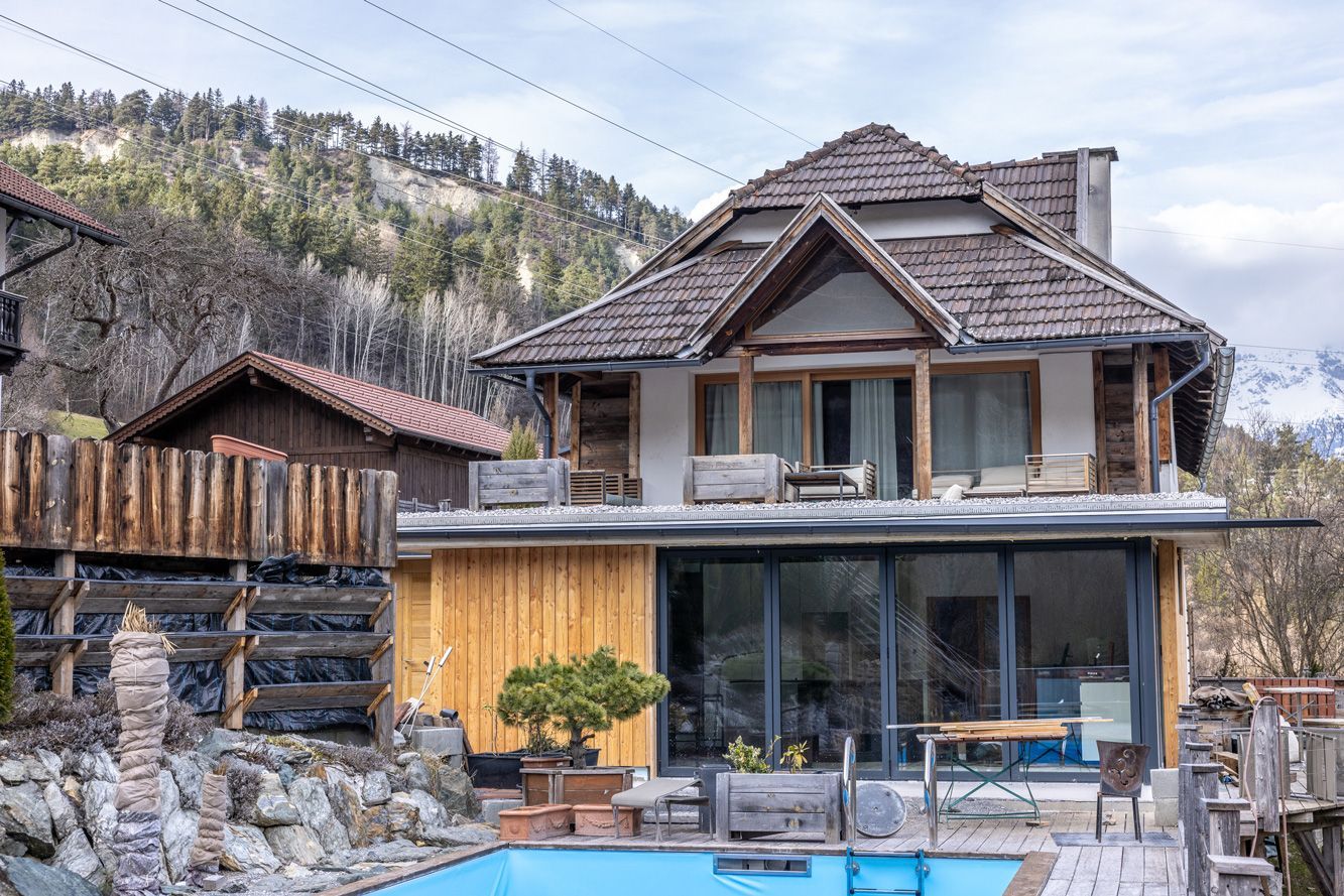 Rustic hillside house with wooden deck and blue pool, set among bare trees and rocky landscape