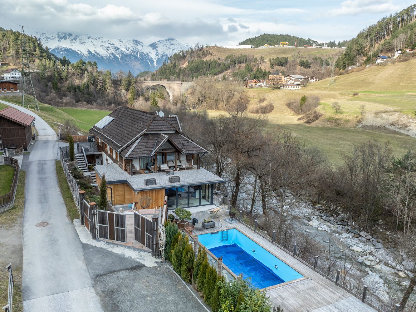 Mountain chalet with a blue pool in a snowy valley, surrounded by hills and a winding road.