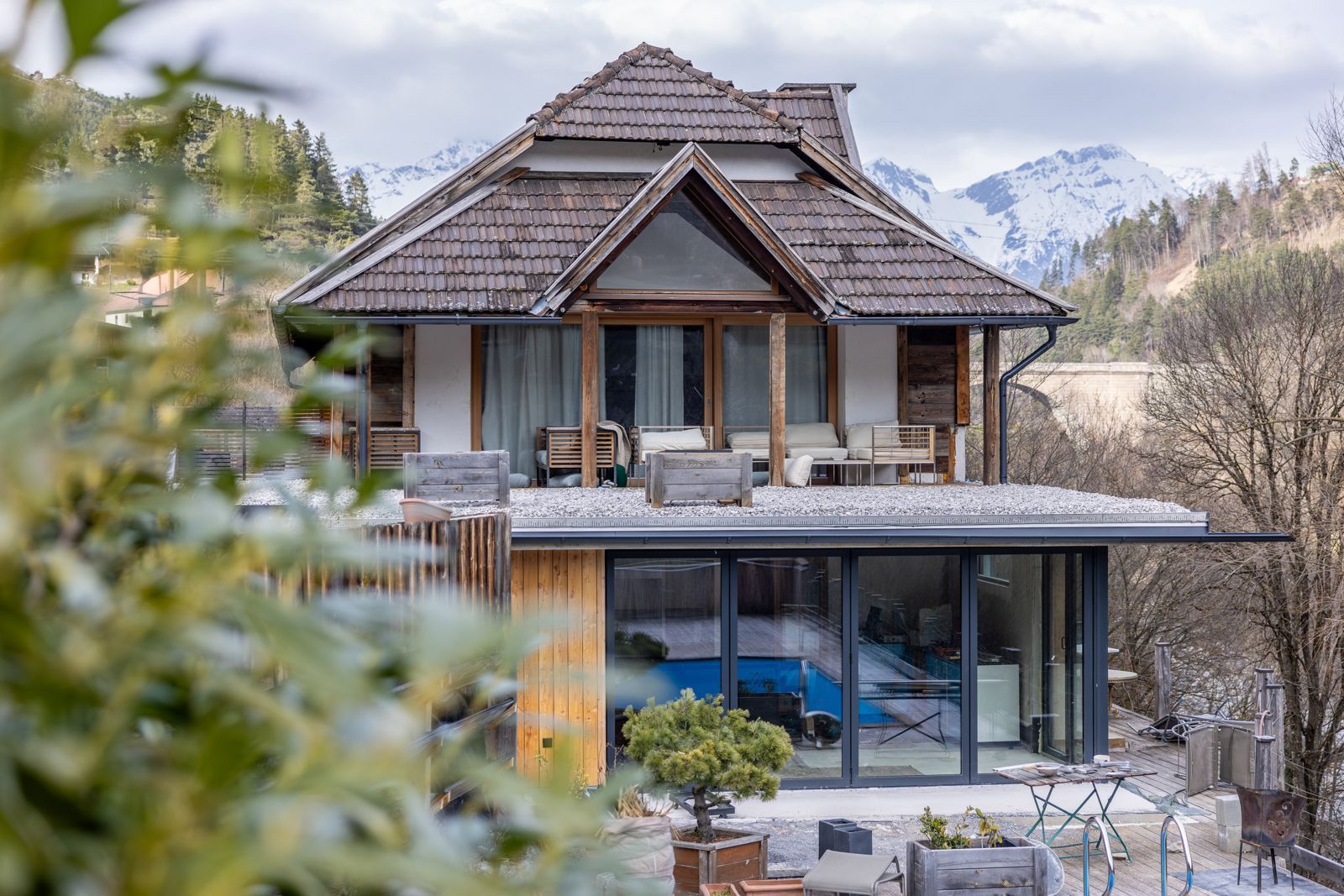 Large modern house with a dark tiled roof and glass lower level, set among trees and mountains