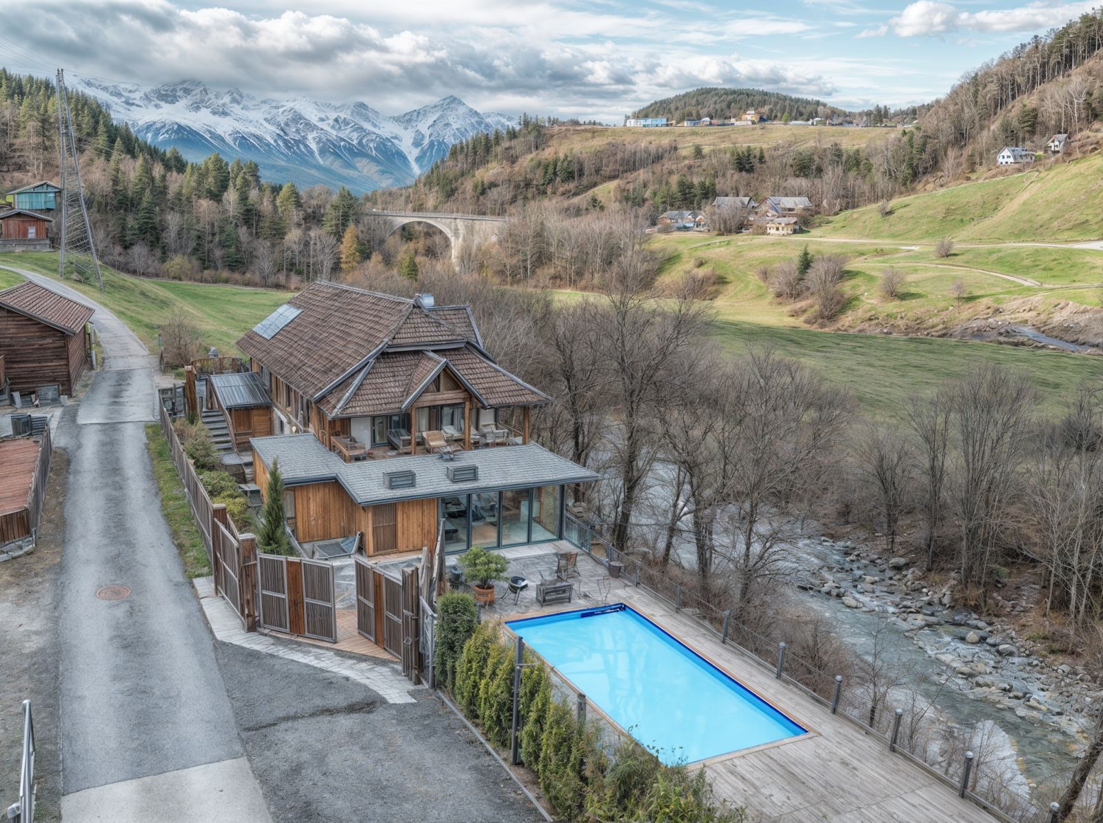 Mountain chalet beside a blue pool, with snowy peaks and a winding road in a rural valley