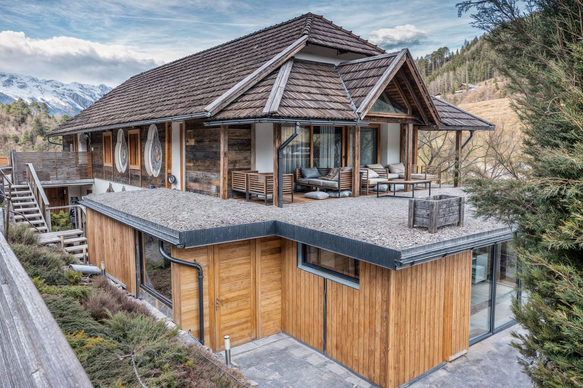 Modern mountain chalet with wood siding, stone roof, and large windows overlooking snowy peaks