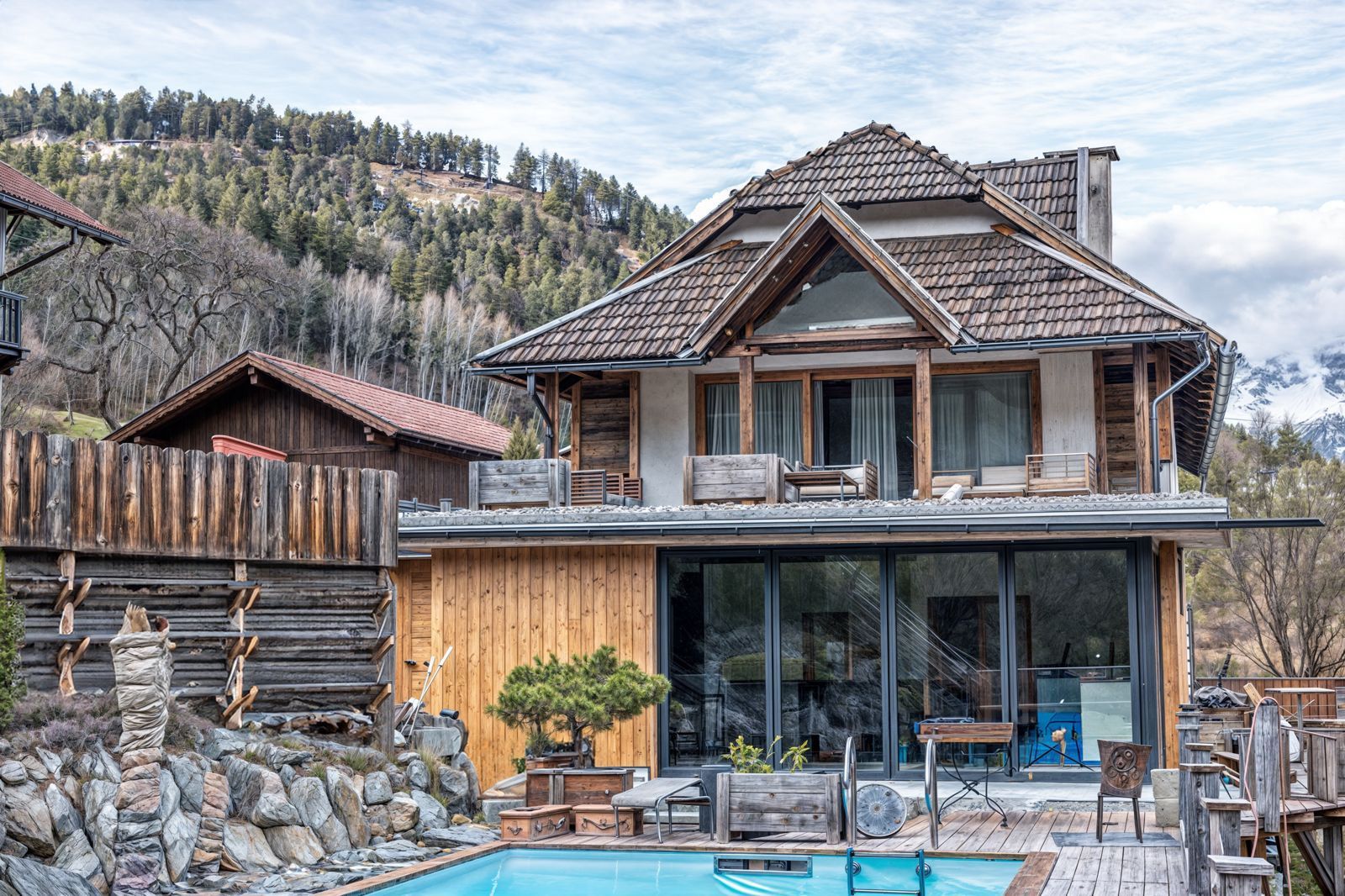 Rustic mountain house with stone and wood exterior beside a blue pool, backed by forested hills