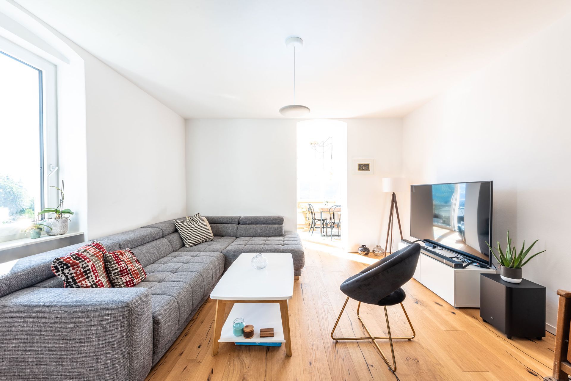 Bright living room with gray sectional sofa, TV, white walls, wooden floor, and large window.
