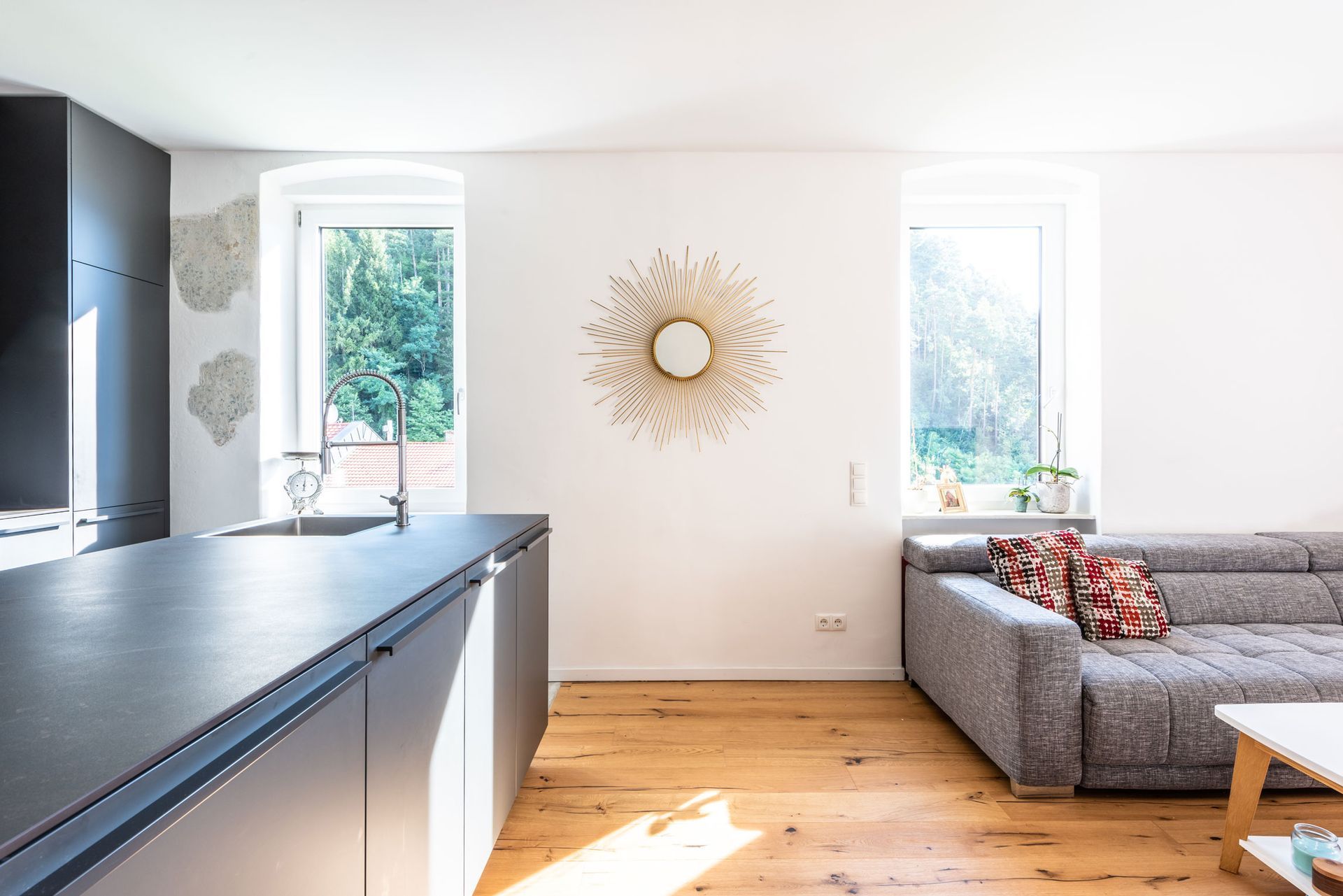 Bright modern living room with gray sofa, sunlit windows, wooden floor, and kitchen counter in foreground