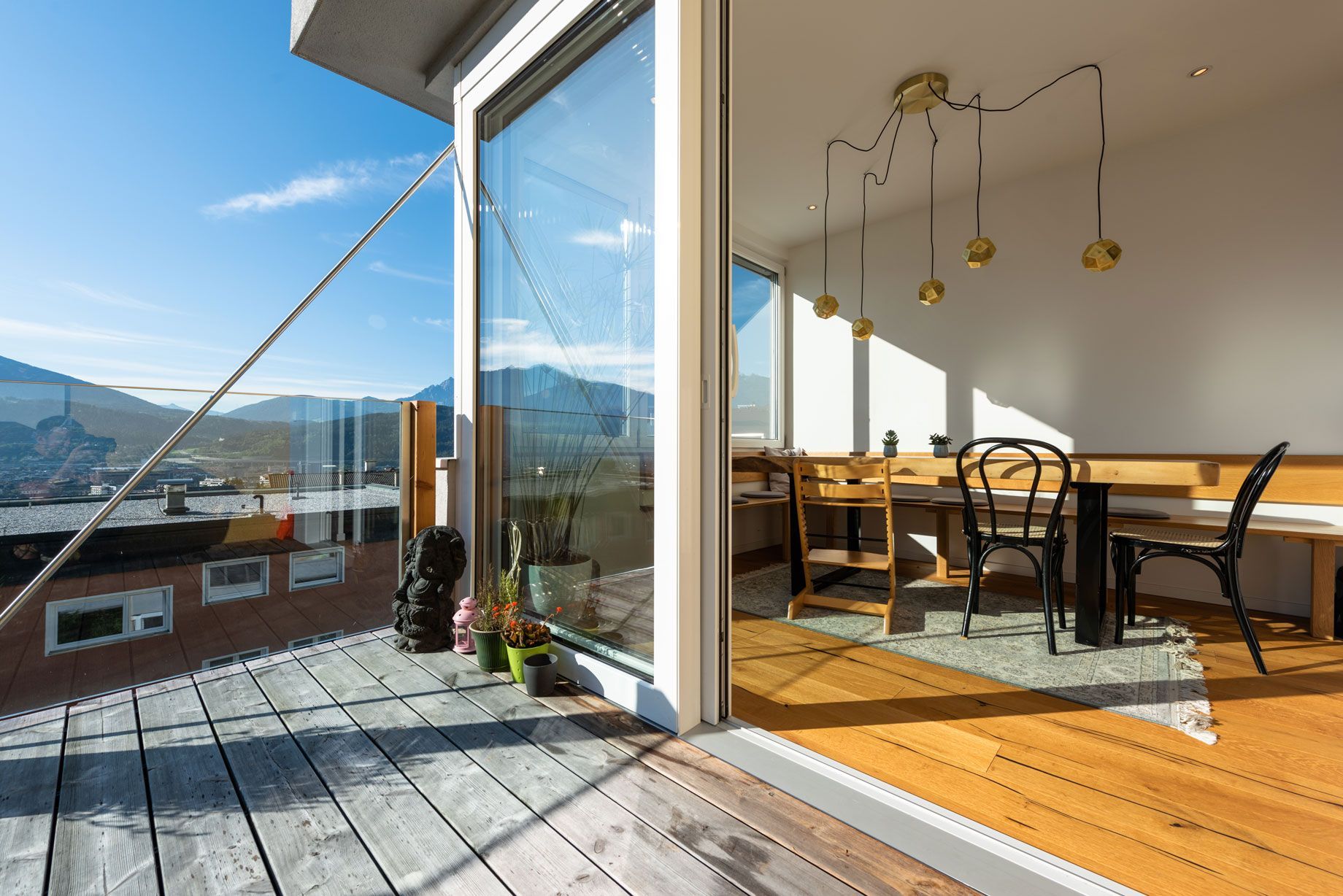 Balcony overlooking mountains, with open glass doors to a bright dining area and wooden table.