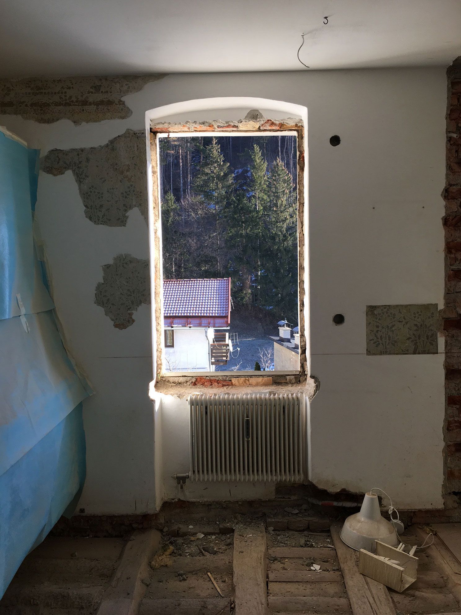 Partially renovated room with peeling walls, window, radiator, and debris on the floor