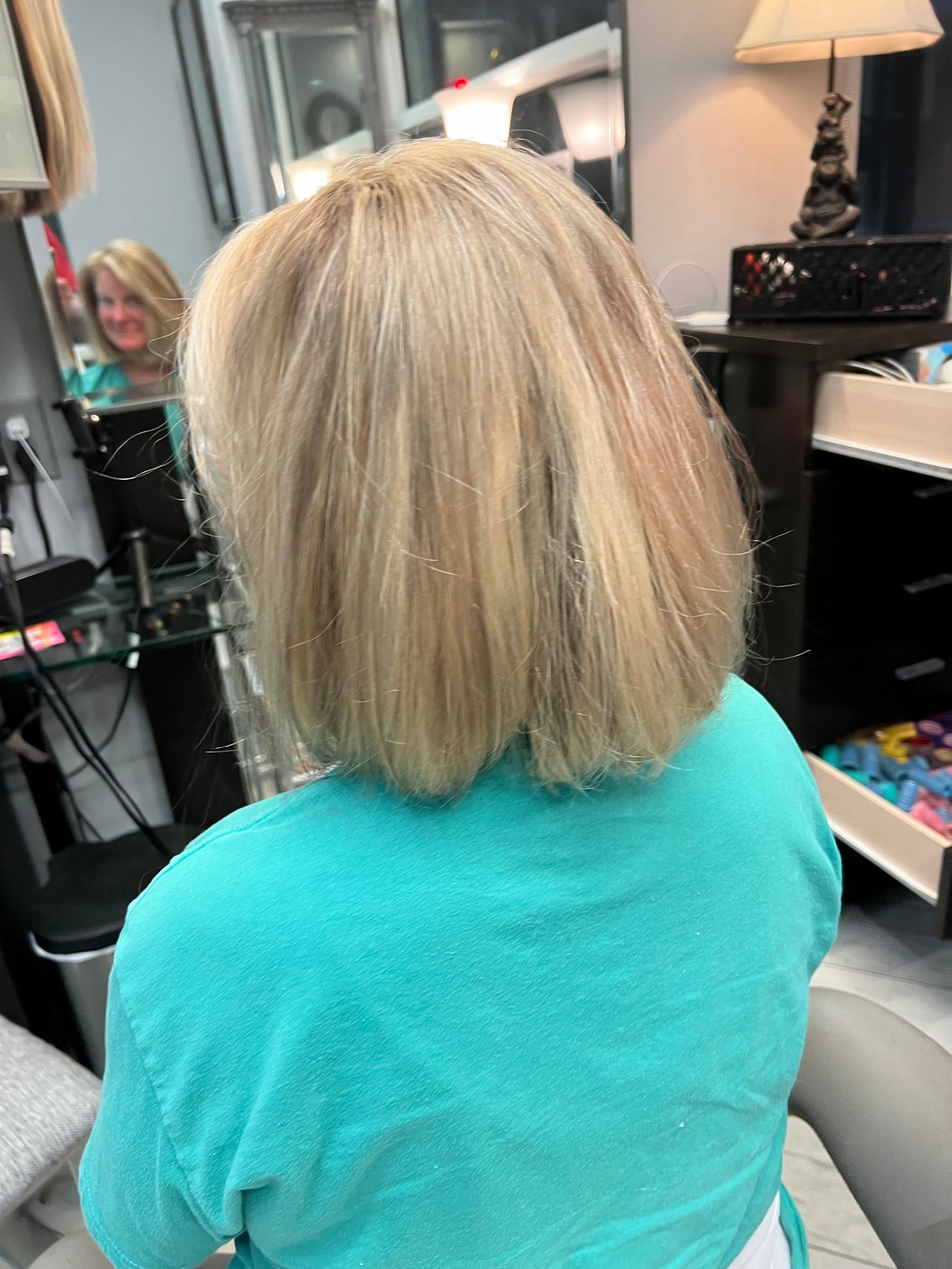 Woman with blonde bob haircut wearing teal top in a salon, looking in the mirror.