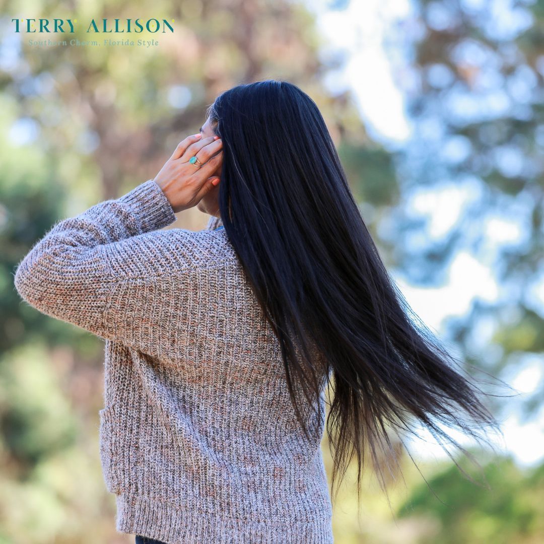 Woman with long black hair wearing a tan sweater, her hand touching her head outdoors.