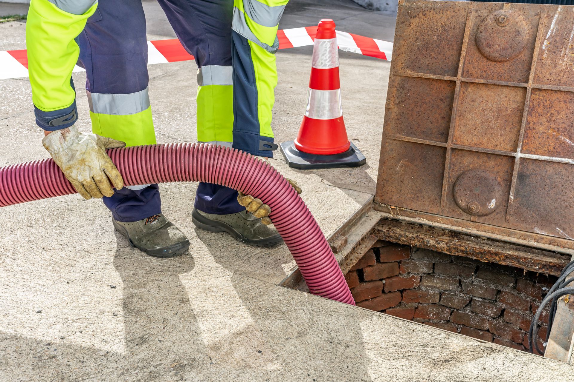 Worker with hose cleaning sewer. Red and white cone, brick interior. Concrete.