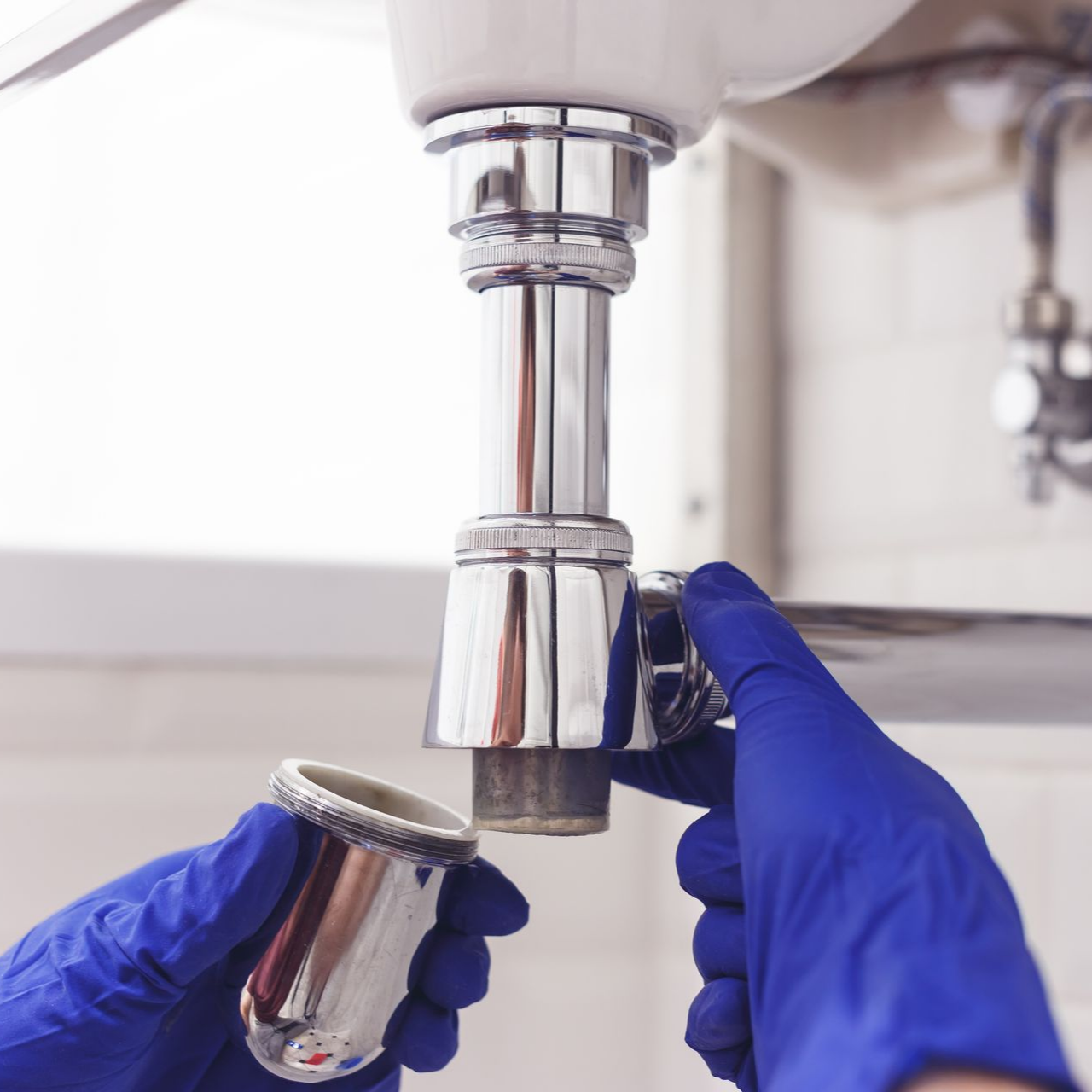 Hands in blue gloves unscrew a drain trap beneath a sink. Hands in blue gloves unscrew a drain trap beneath a sink.