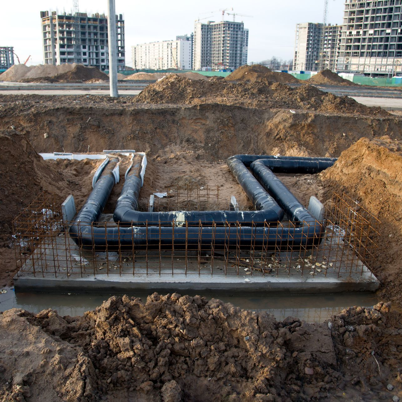 Underground water pipes in a construction site, with dirt mounds and unfinished buildings in the background. Underground water pipes in a construction site, with dirt mounds and unfinished buildings in the background.