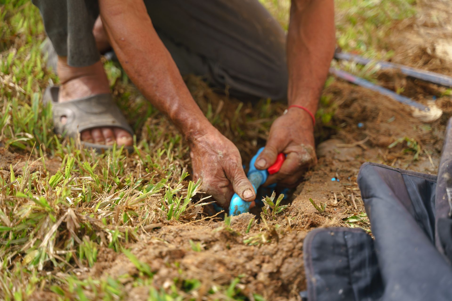 Person kneels in dirt, connecting blue pipes in a grassy area. Hands work on plumbing.