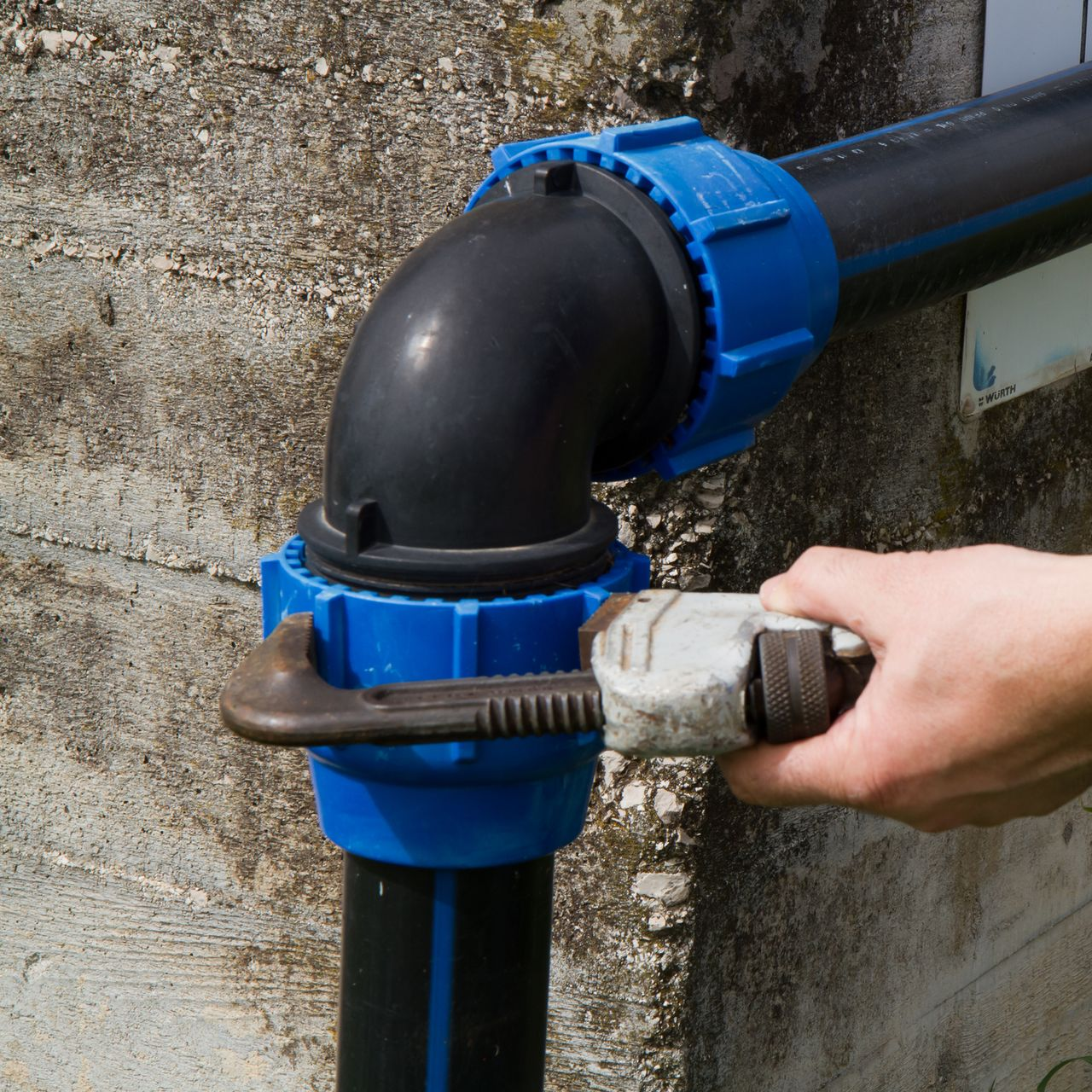 Hand using a wrench to tighten a blue plastic pipe fitting on black pipe near a wall. Hand using a wrench to tighten a blue plastic pipe fitting on black pipe near a wall.