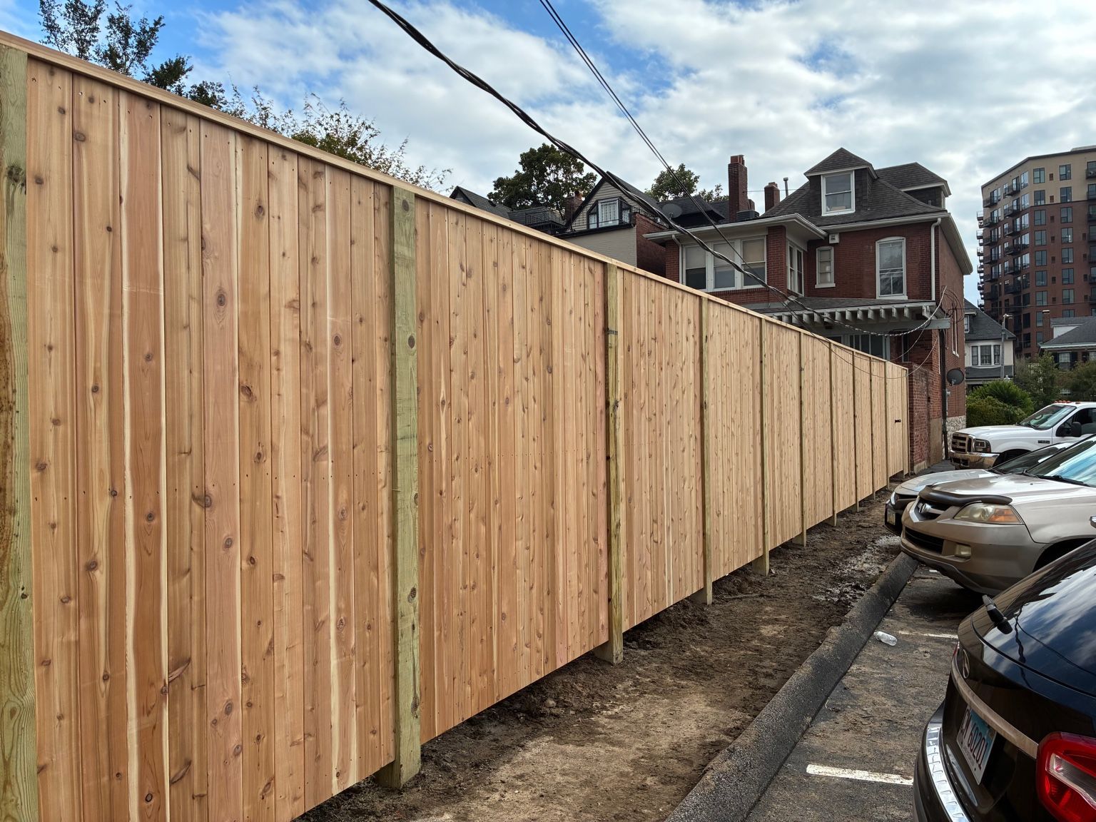 A long wooden fence bordering a parking lot. Buildings and cars are visible in the background.