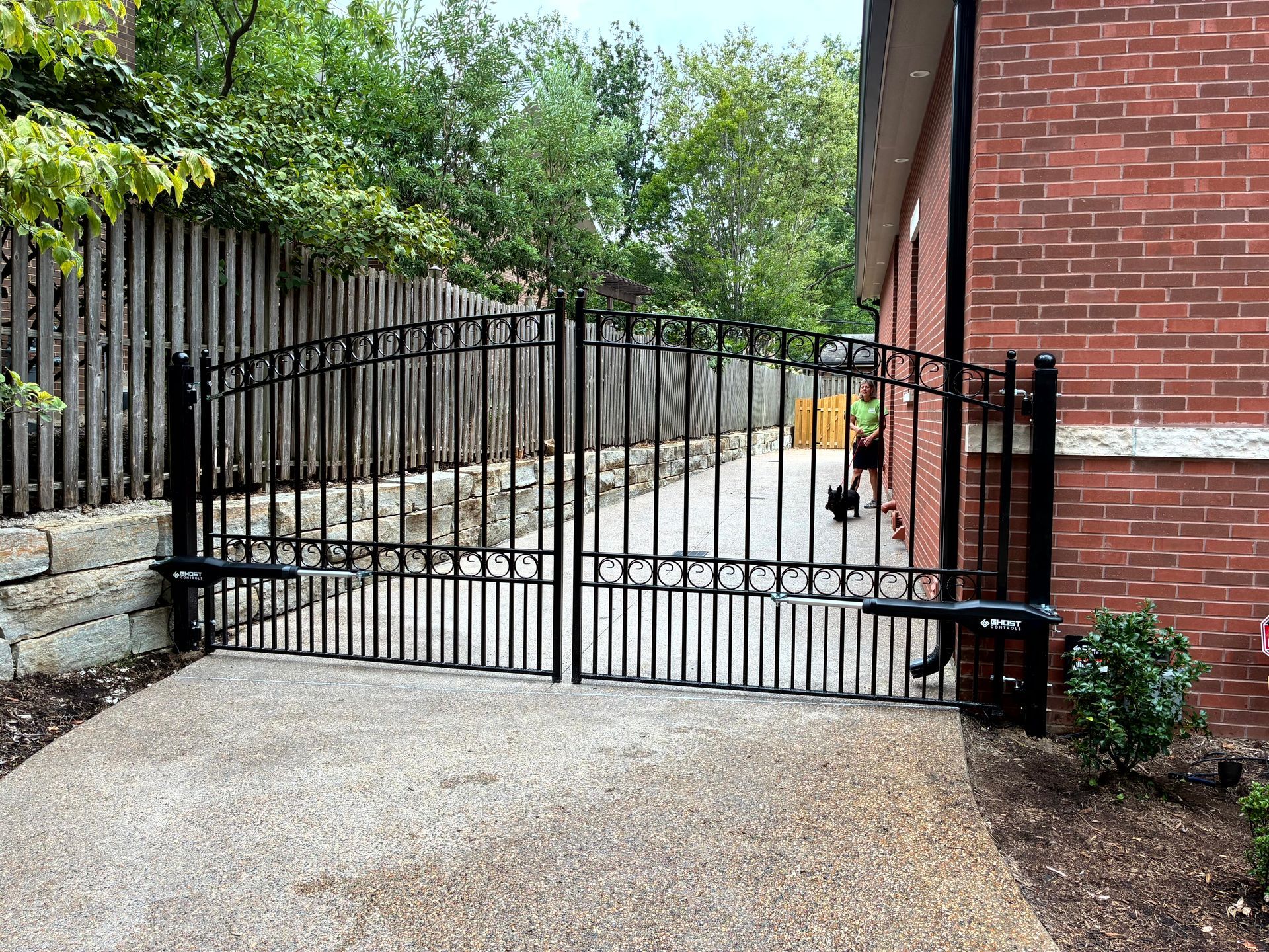 Black wrought iron driveway gate on concrete, next to a brick building and wooden fence.