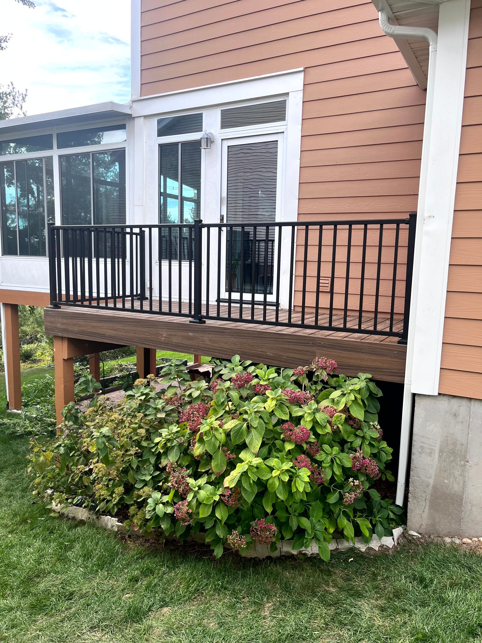 Deck with black railing, door, and bushes. The house has wood siding and a covered porch.