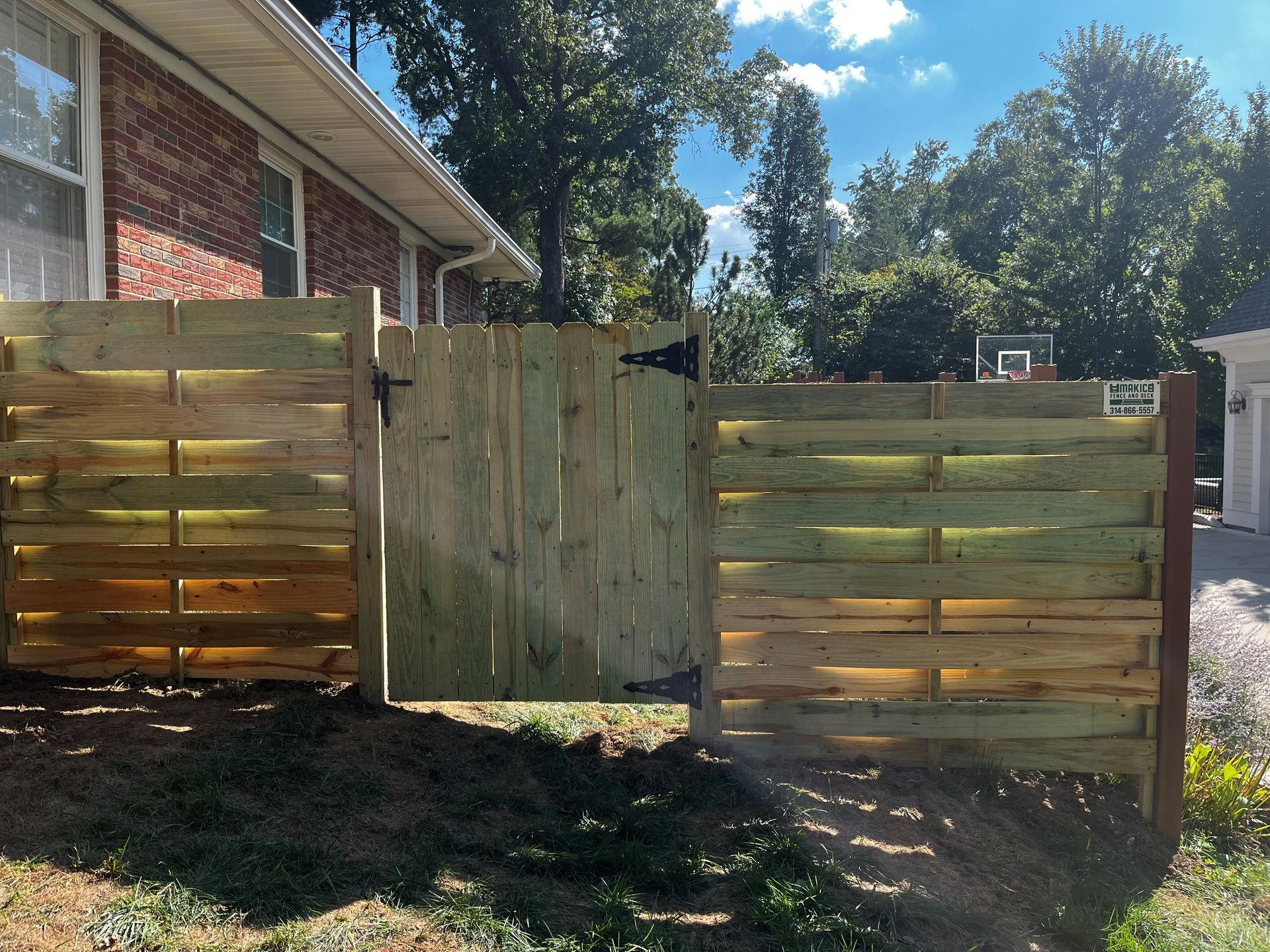 A wooden fence with a gate in front of a brick house. Green grass and trees are in the background.