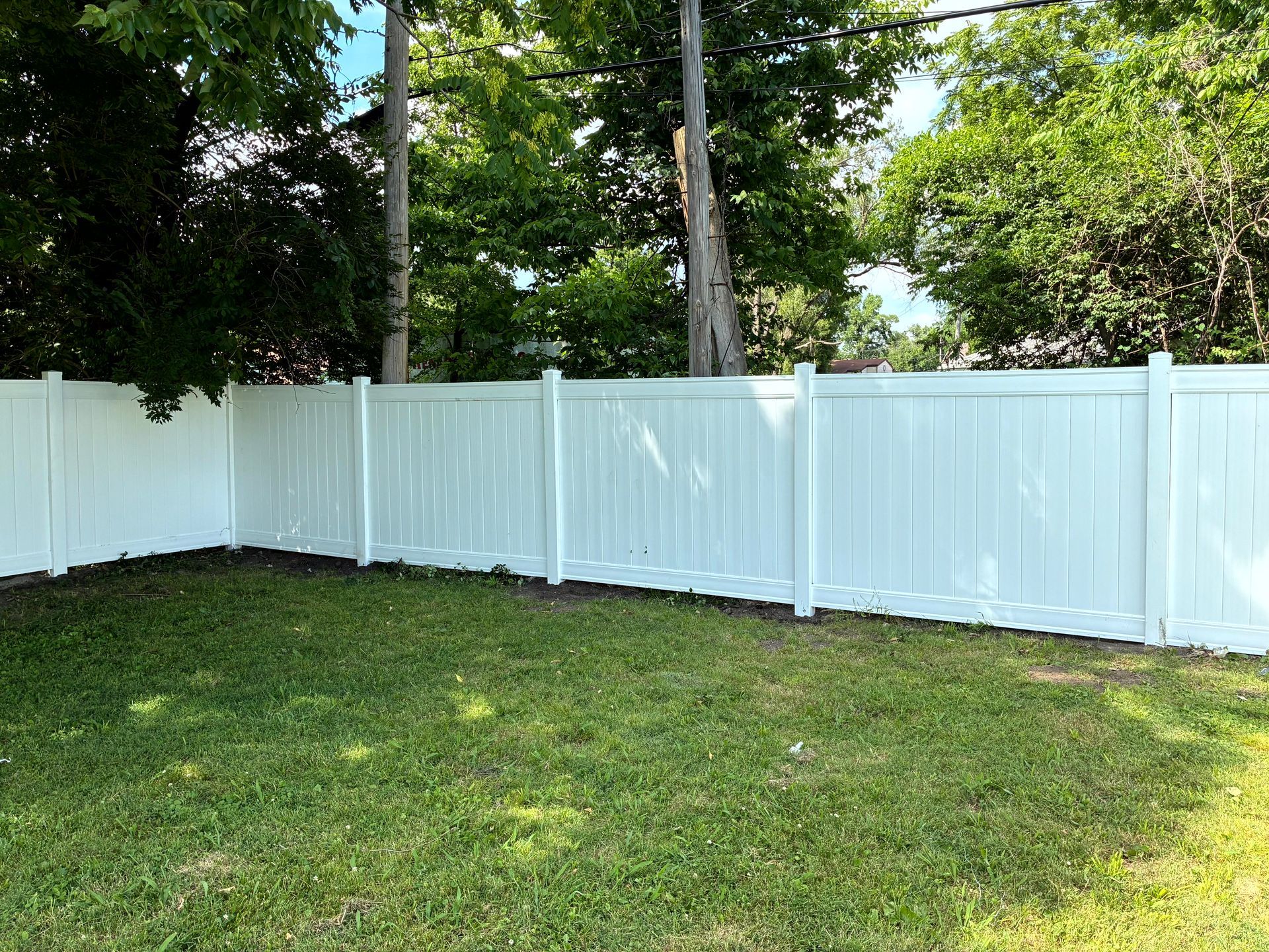 White vinyl fence encloses a green grassy yard, surrounded by trees under a blue sky.