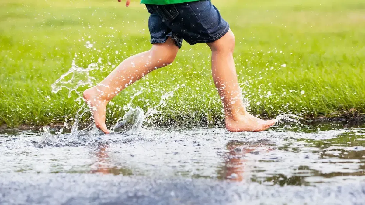 Child running through a shallow puddle on green grass, splashing water with bare feet