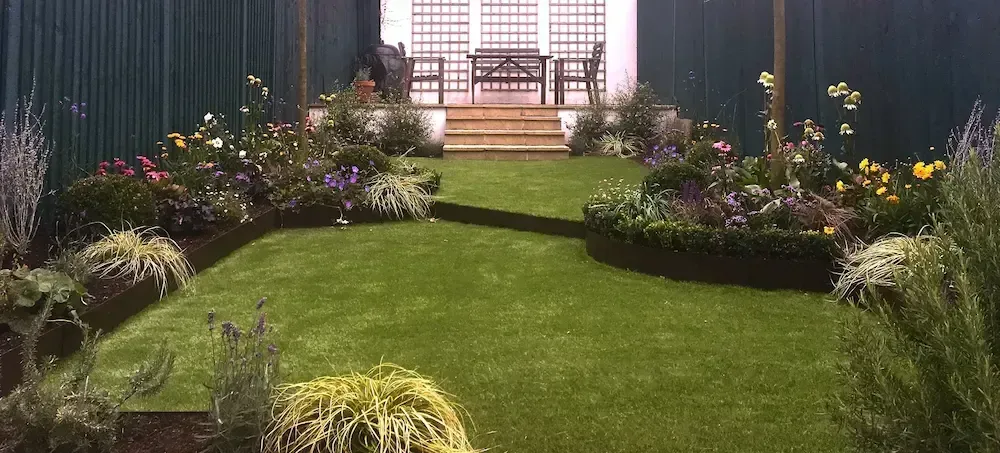 Lush garden path leading to steps and a white pergola, with flower beds and a green lawn on both sides