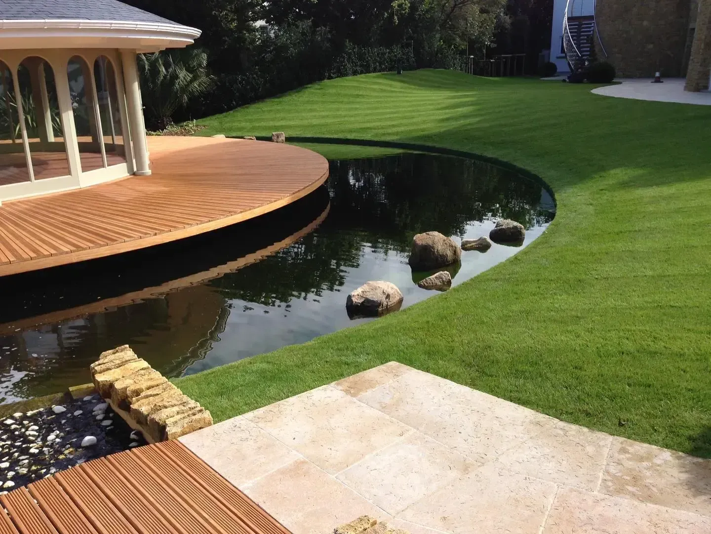 Curved wooden deck beside a small pond and green lawn in a landscaped backyard