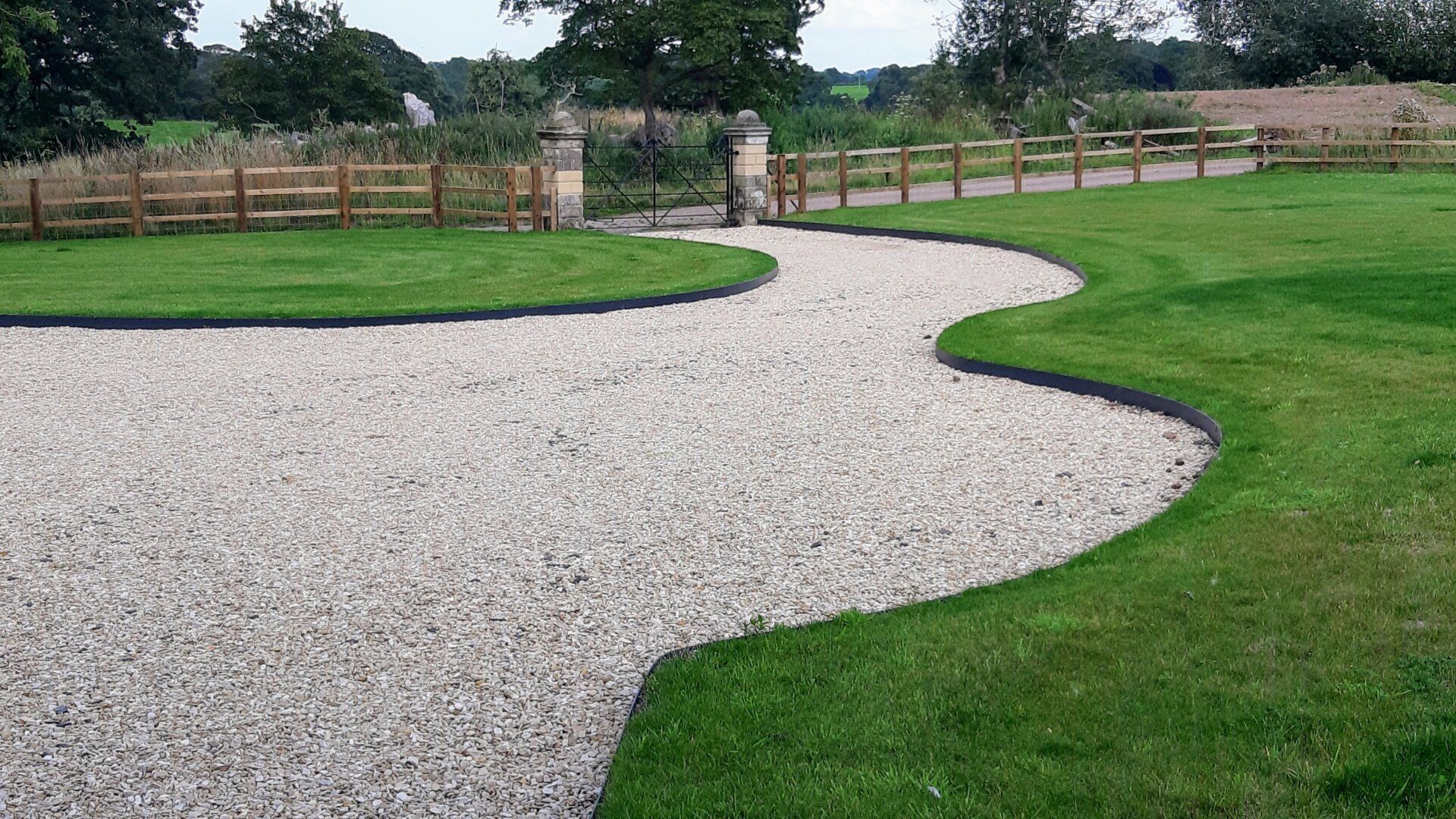 Curved gravel path through green lawn, leading to a fenced garden gate under cloudy sky.