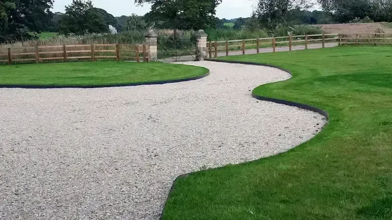 Curving white gravel path through a green park, bordered by grass and a wooden fence.