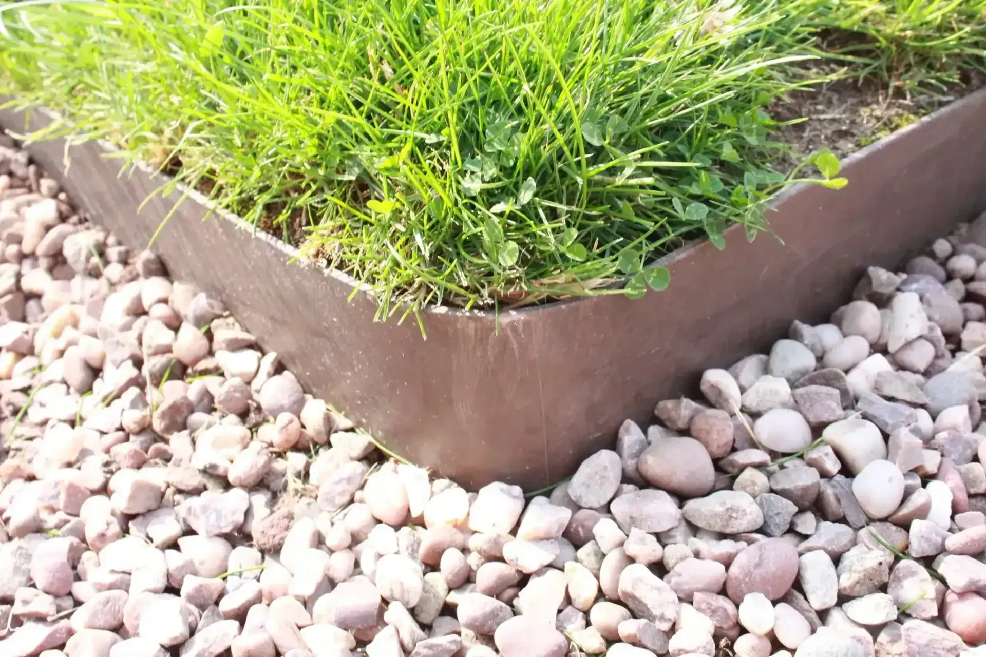 Corner garden bed with green grass bordered by brown edging and white pebbles