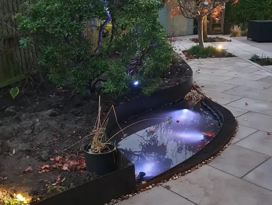 Garden path beside a small illuminated pond at dusk, with reflections and surrounding plants