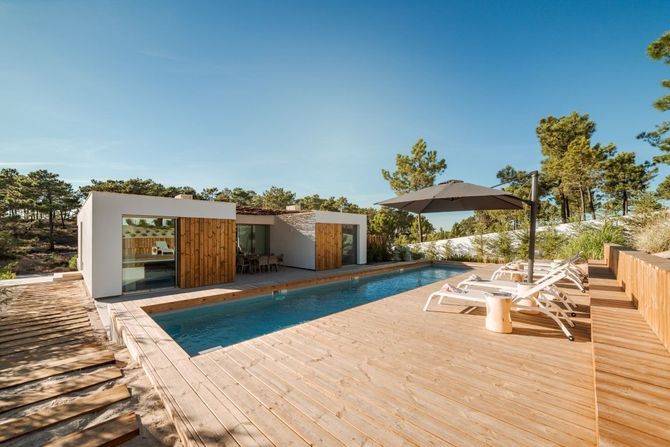 Modern villa with wooden deck and pool, surrounded by trees and lounge chairs under a blue sky