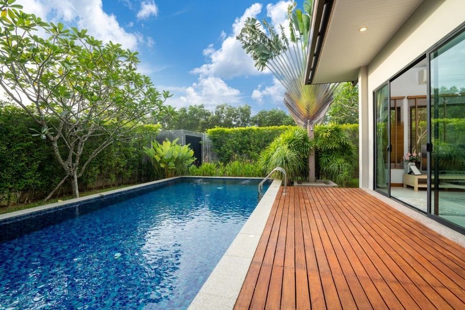 Modern backyard with a blue pool, wooden deck, and glass-walled house under a sunny sky