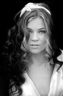 A black and white photo of a woman with long curly two-toned hair wearing a headband and a white dress.
