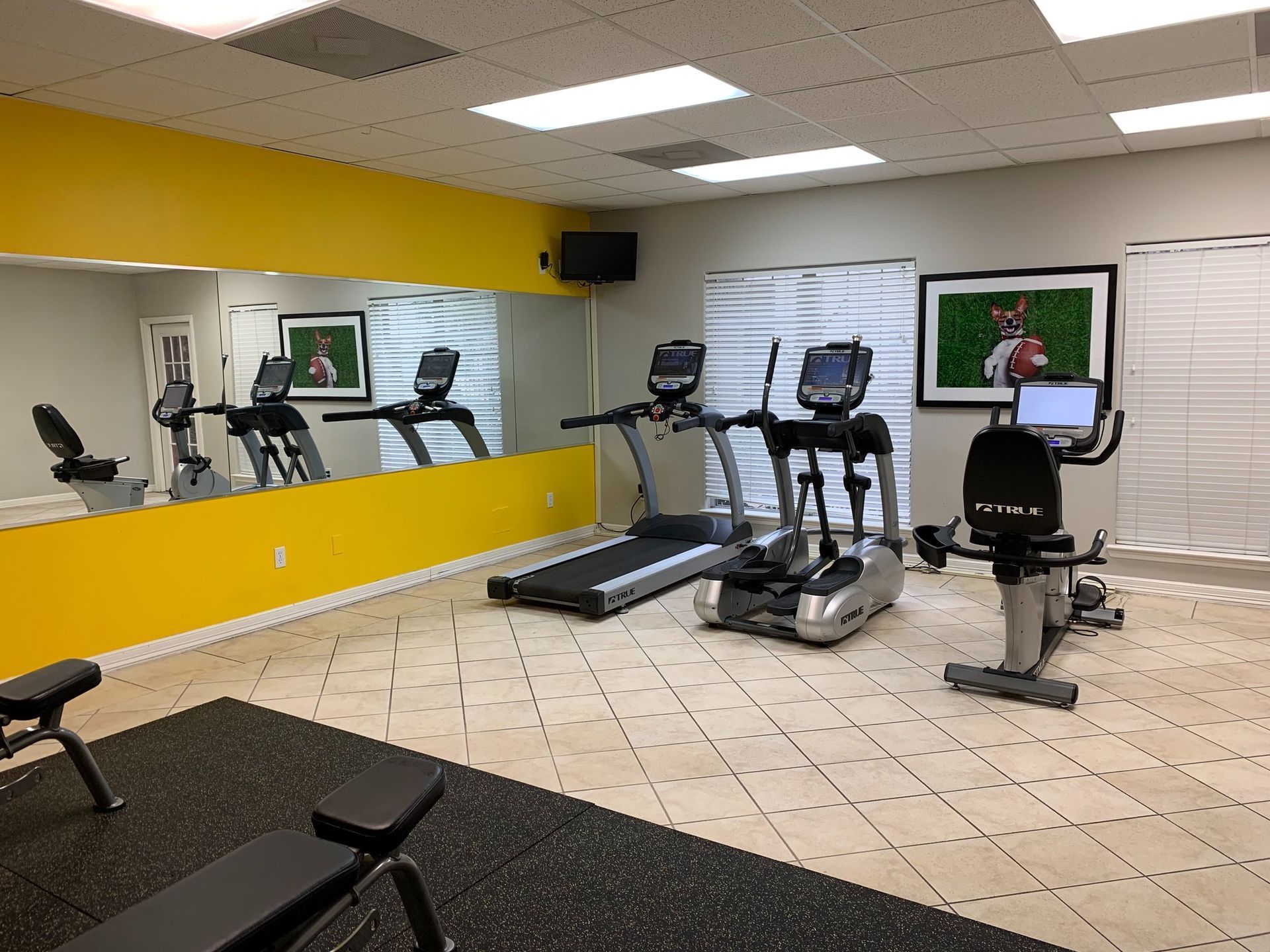 Small gym with treadmills, ellipticals, and a stationary bike on tiled floor, mirror wall and yellow accent wall