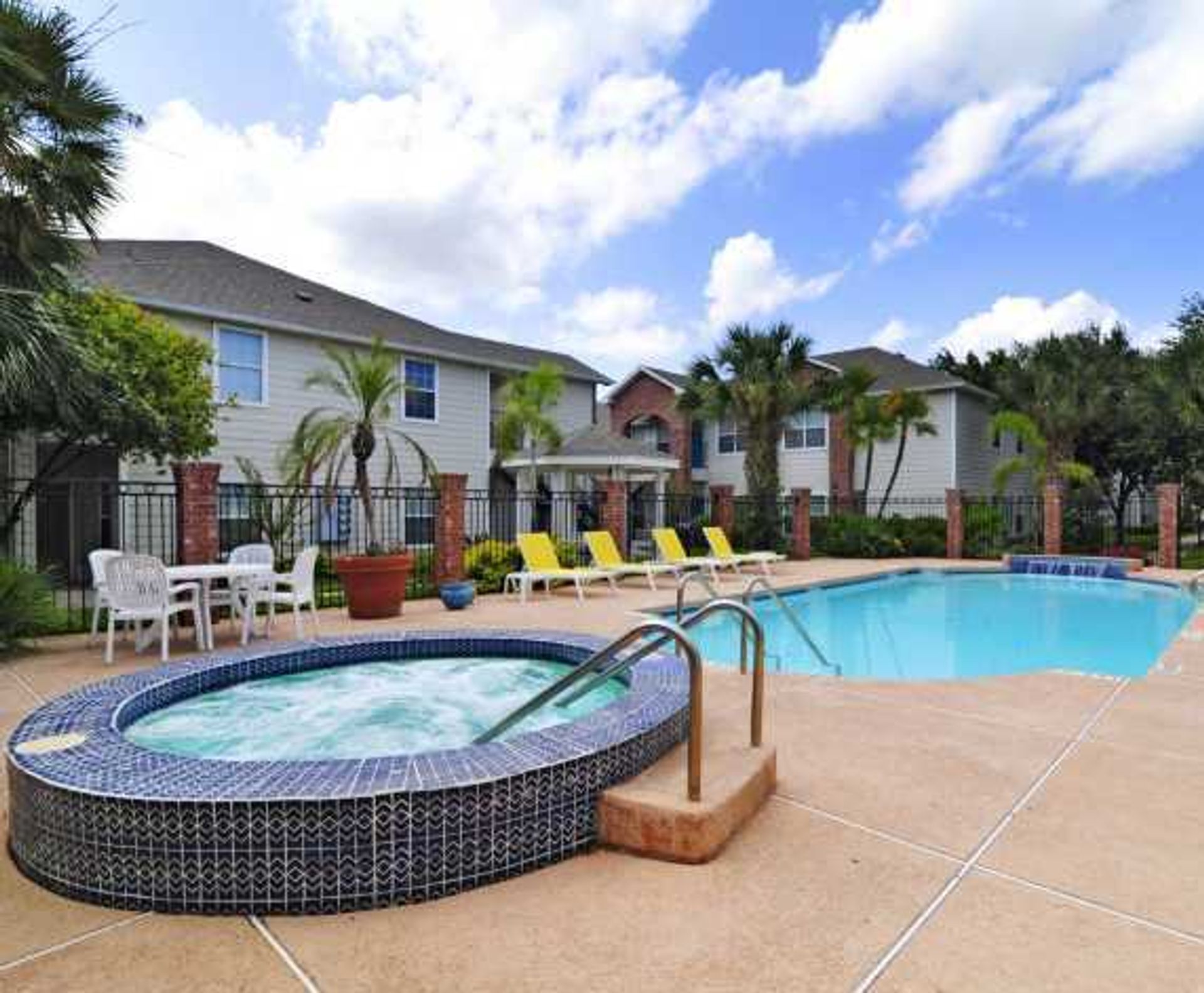 Resort-style pool with hot tub, lounge chairs, and palm trees beside apartment buildings